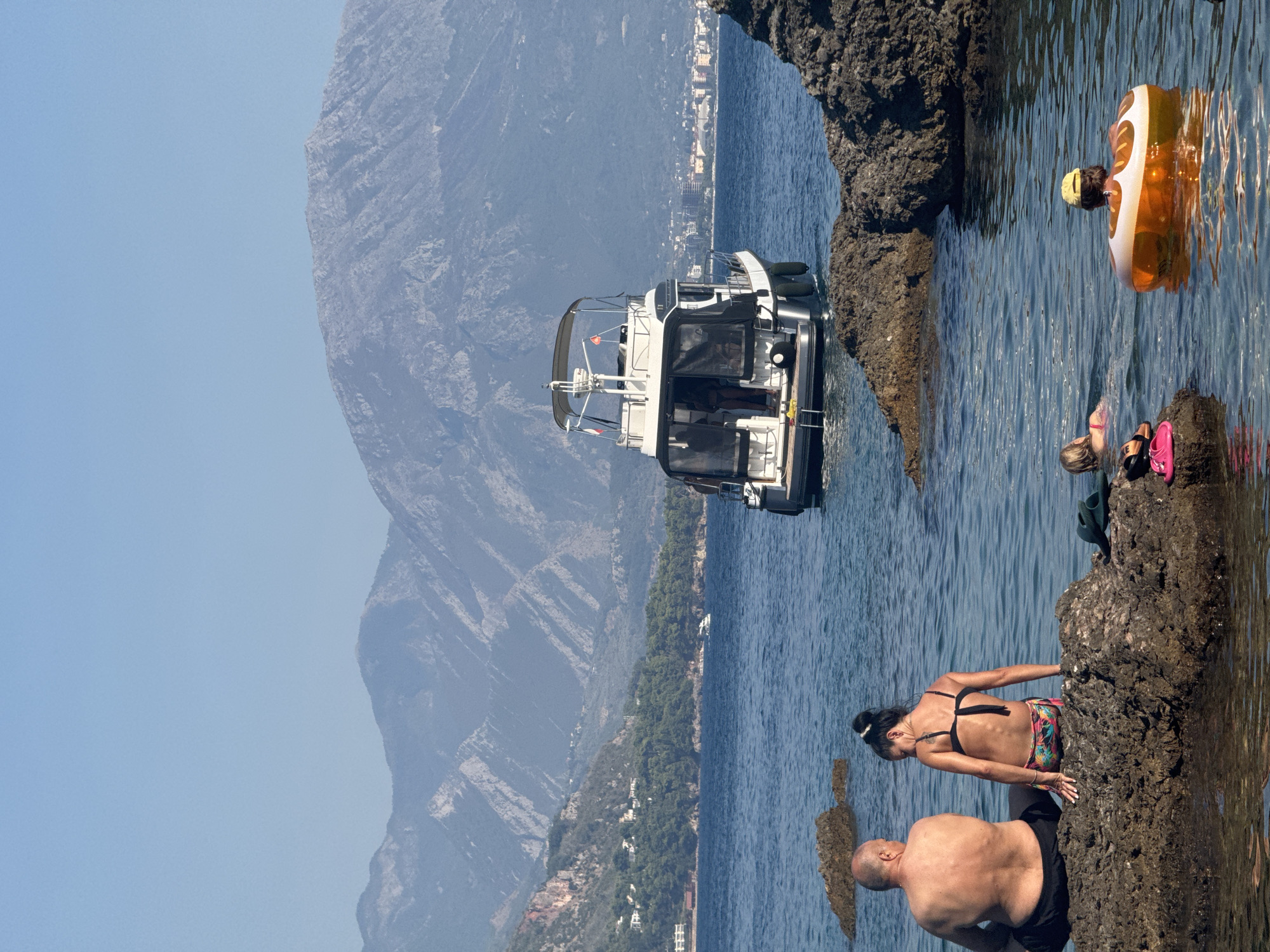 In Sutomore, Montenegro, a serene coastal scene unfolds under a clear blue sky. In the foreground, two individuals, a shirtless man with dark hair and a woman with dark hair in a bikini, sit on a rock at the edge of the water. They face away from the camera, gazing out at the calm, blue sea. Further out in the water, a white motor yacht with black accents sails away from the shore. In the middle ground, a small boat can be seen near a cluster of trees and buildings on a hillside. In the background, imposing mountains rise, their rugged slopes covered in sparse vegetation and dotted with buildings. The scene is peaceful, with the gentle lapping of waves and the distant hum of the boat motor creating a tranquil atmosphere.