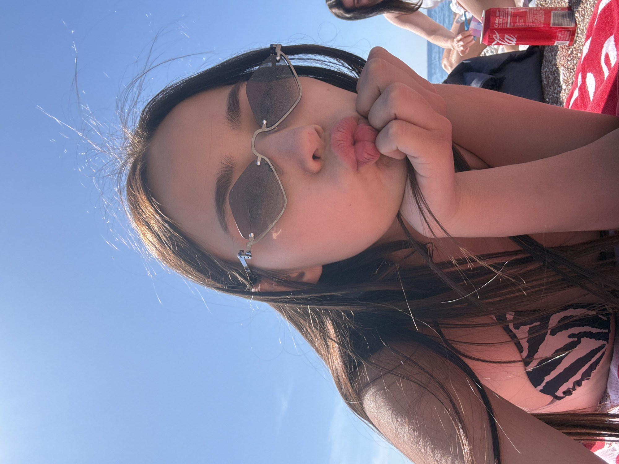 A young woman with long brown hair is on a beach, wearing sunglasses and a zebra-striped swimsuit. She is making a kissing face and holding her cheeks with her hand. The sky is blue. A red and white towel and a can of Coca-Cola are visible in the background, along with other people. The location is Unknown, Unknown.