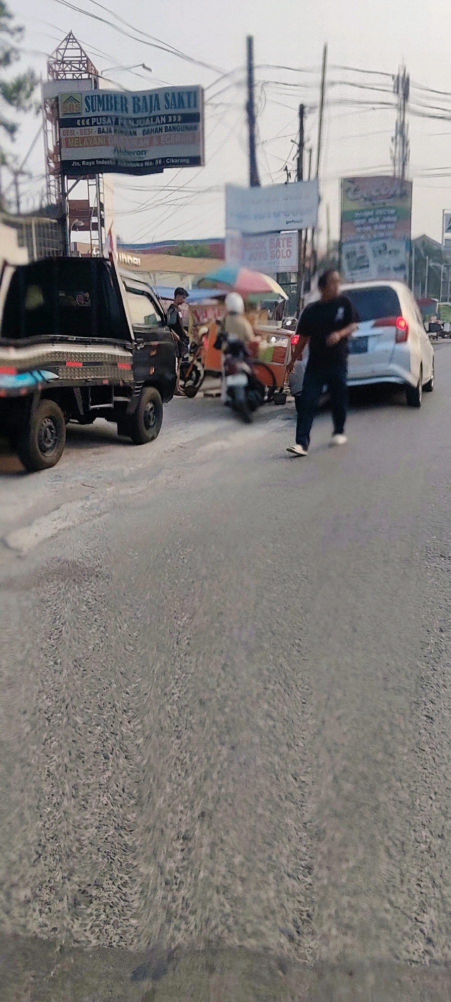 In Mekarmukti, Indonesia, on a street, a black pickup truck is parked. To the right of the pickup truck, a person in a black t-shirt and dark pants walks across the street. A silver van is parked on the right side of the street. A food cart is parked on the left side of the street, with a person on a motorcycle next to it. Another person is walking near the food cart. Overhead, there are power lines and a street sign. The ground is asphalt.