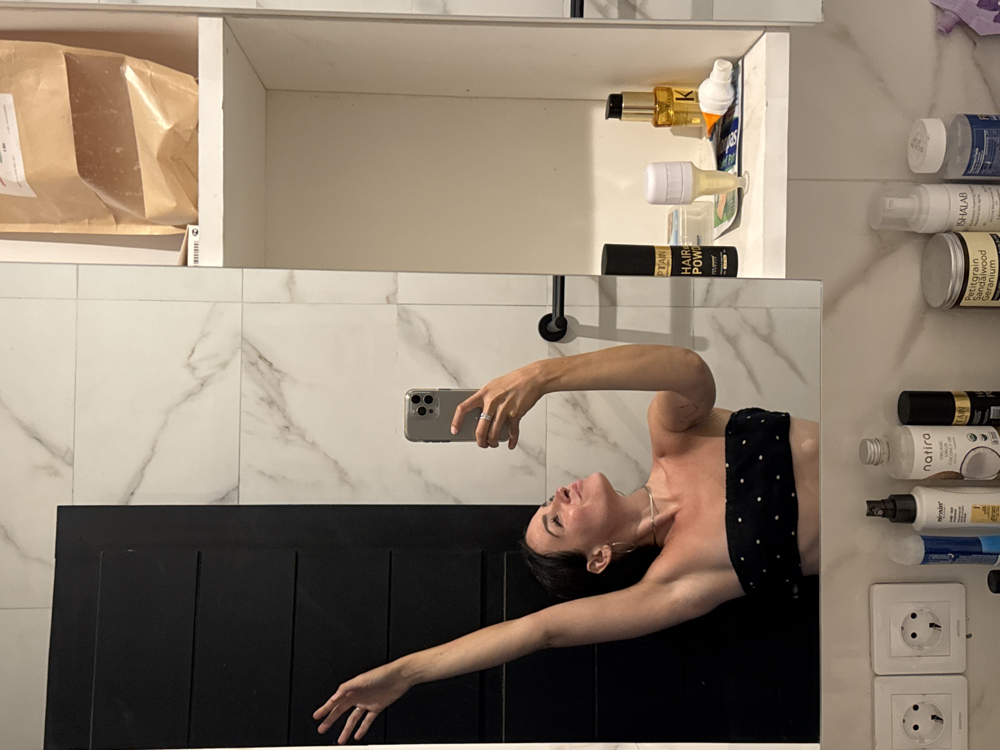 This media file shows a person taking a selfie in a bathroom. The person is a young woman with dark hair, wearing a black and white polka dot top and a silver necklace. She is lying down and reaching her arm up to take the photo with her phone. The bathroom has white marble tiles on the walls and a black wooden cabinet below. On a shelf above, there are various toiletries and beauty products, including bottles of shampoo, conditioner, and hair powder. Some of the visible text on the products includes "nioxin," "natira," "ORGANIC VIRGIN COCONUT OIL," "Petitgrain Sandalwood Geranium," and "KERASIS." The lighting suggests it is daytime, likely indoors. The overall mood appears casual and relaxed.
