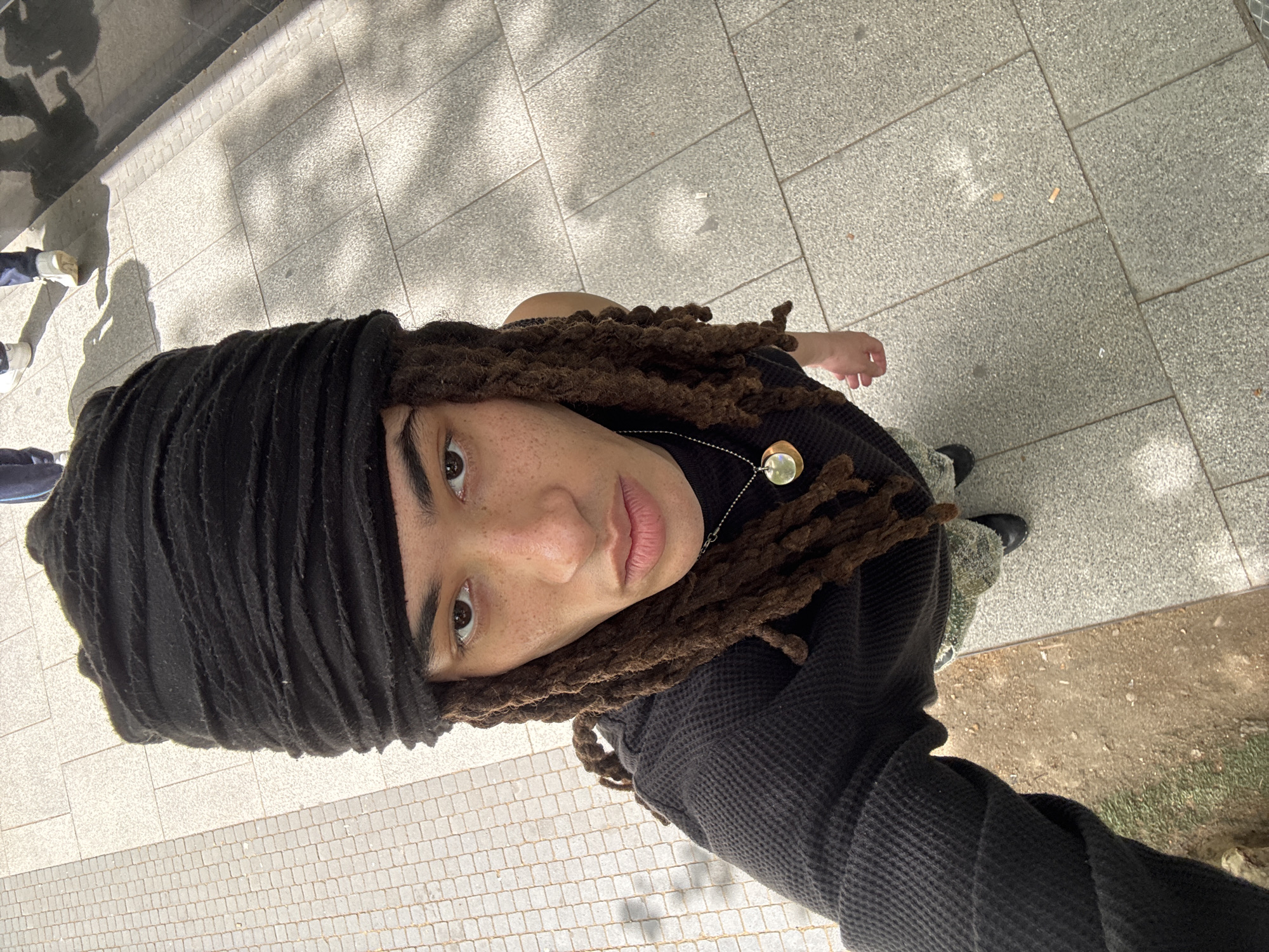 An overhead photograph captures a person looking directly up at the camera in an urban outdoor setting in Madrid, Spain. The individual has brown dreadlocks, partially covered by a black, textured headwrap or scarf. Their face shows visible freckles across the nose and cheeks, and they have light brown eyes and neutral-toned lips. A silver chain necklace with a circular, dual-toned metallic pendant (one side appearing gold, the other silver and reflective) rests on their chest. They are wearing a dark, possibly black, textured garment, resembling a waffle-knit fabric, and a black glove or sleeve on their visible left arm, which extends towards the bottom right of the frame.
The ground is paved with light grey, rectangular flagstones or tiles, characteristic of city pedestrian areas. Strong, defined shadows of trees or other structures, along with shadows of other people's legs and feet, are cast across the pavement, indicating bright sunlight and daytime. In some areas, scattered debris including two cigarette butts are visible on the paving. A narrow strip of dry earth and sparse green grass can be seen at the far right edge of the frame, bordering the paved area. The person is stationary, posing for the photograph. There is no visible text.