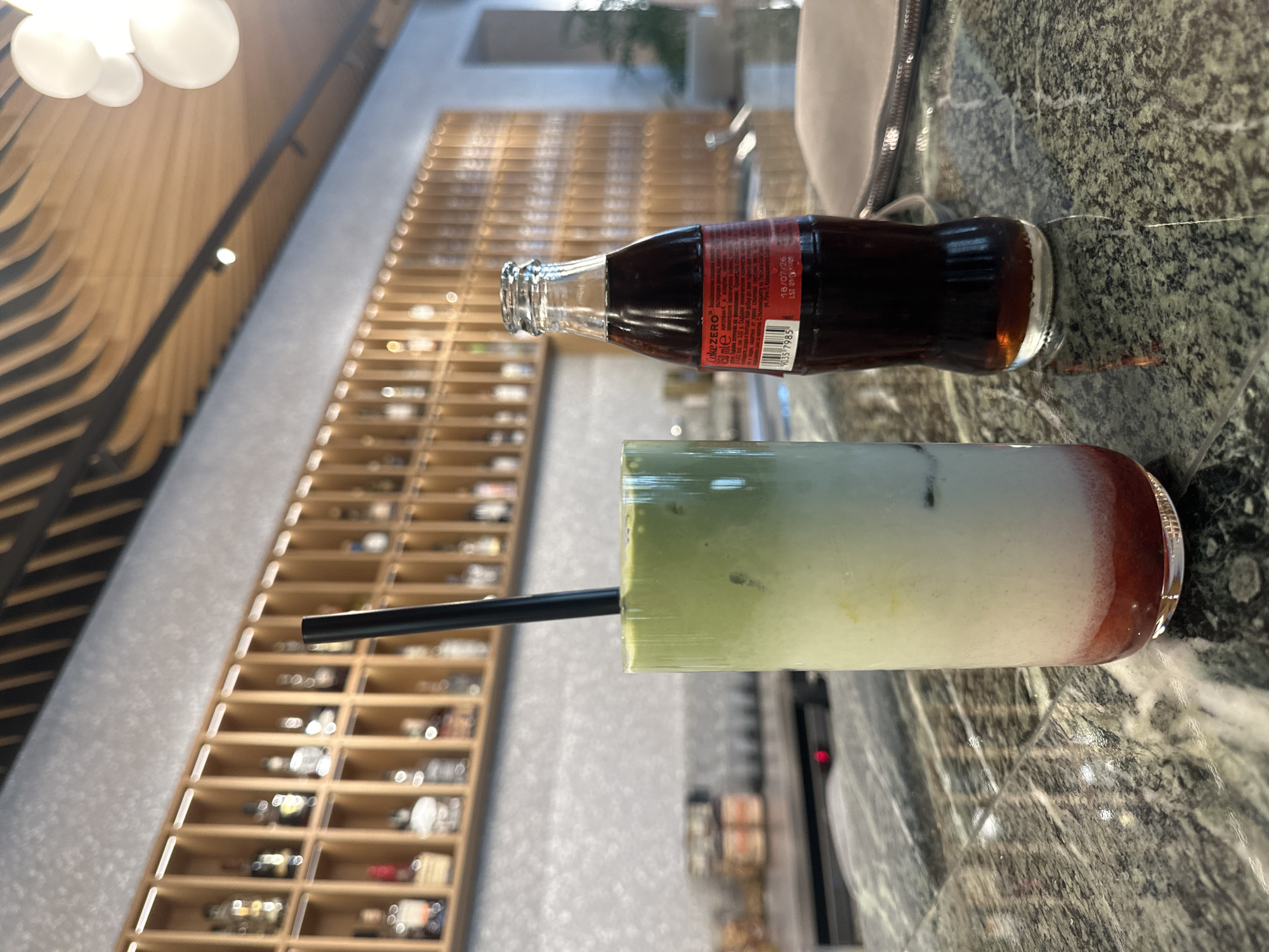 The image displays an interior bar scene, likely situated in Varna, Bulgaria, focusing on a dark green marble-patterned countertop. On the left, a tall, clear glass contains a layered beverage, visibly composed of a red liquid at the bottom, a white milky substance in the middle, and a green layer on top, with ice cubes interspersed throughout; a black straw is inserted into the drink. To the right of the glass, a 250ml glass bottle of Coca-Cola Zero stands, identifiable by its red label with white text including "COKE ZERO," "250 ml e," Cyrillic script, and a visible date of "18/07/26." A light-colored zippered pouch is partially visible on the far right of the counter.
The background consists of a light gray, textured wall, above which a long wooden back bar shelving unit is mounted. This unit features numerous individual compartments, each holding a blurred bottle, consistent with liquor or spirit bottles. The ceiling above is characterized by an architectural design of wavy, undulating wooden slats and incorporates several spherical white light fixtures, alongside a black track light with a single spotlight. Farther into the background on the right, a blurred arched doorway or opening is visible. No individuals are present in the frame.