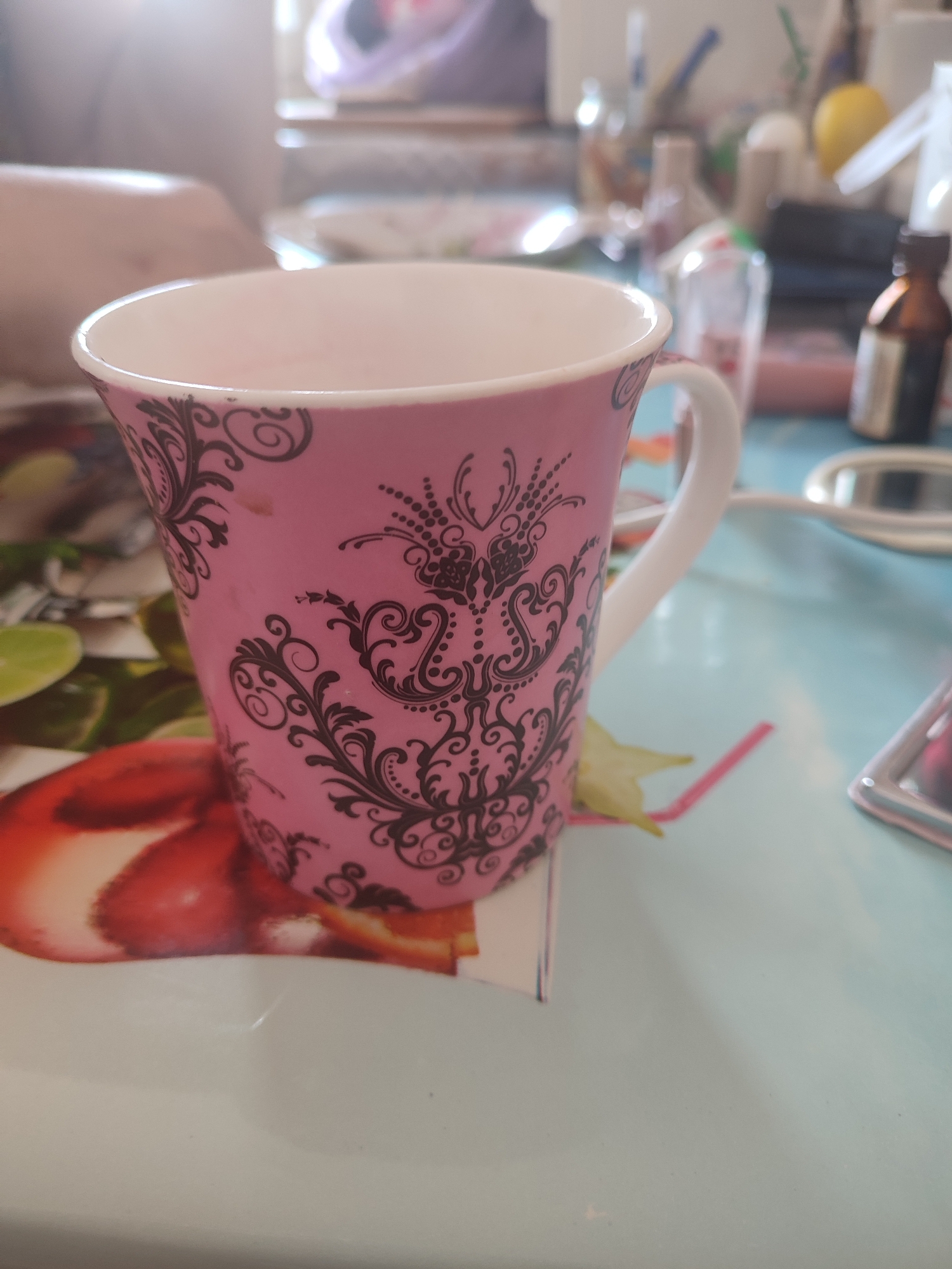 The image shows a pink mug with black ornate patterns on a light blue table. The mug is positioned on the right side of the frame and is partially filled with a light-colored liquid. In the background, out of focus, there are various objects, including bottles, a mirror, and possibly some shelving. A person's arm and hand are visible on the left side of the image, resting on the table. A pink straw is also visible in front of the mug. The overall lighting suggests an indoor setting, possibly a home or cafe. The image is not specific to Chernivtsi, Ukraine, and provides no contextual clues to suggest this location.