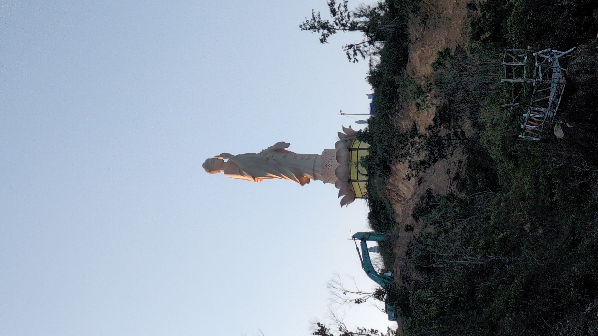A large, light-colored standing Buddha statue is positioned on a vegetated hillside in East Quy Nhon Ward, Vietnam. The statue, illuminated by golden light, stands on a multi-tiered lotus pedestal with a yellow lower band. Its right hand is raised with the palm outward, and its left hand holds drapery. To the left of the main statue, a green excavator is partially visible on the exposed earthy terrain of the hillside. Several smaller white standing statues are discernible near the base of the primary figure. Dense green foliage covers much of the hillside, with some bare branches visible. In the lower right foreground, a weathered wooden structure, resembling an observation platform, is present amidst the vegetation. The sky above is clear and light blue.