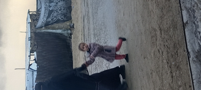 A young child wearing a checkered coat and pink pants is running outdoors, with a dark object in the foreground possibly held by them. Behind the child, the ground appears to be a dirt or gravel surface. In the background to the left, there is a structure covered with fabric or tarps, resembling a makeshift dwelling. The overall scene suggests a setting of poverty or displacement.