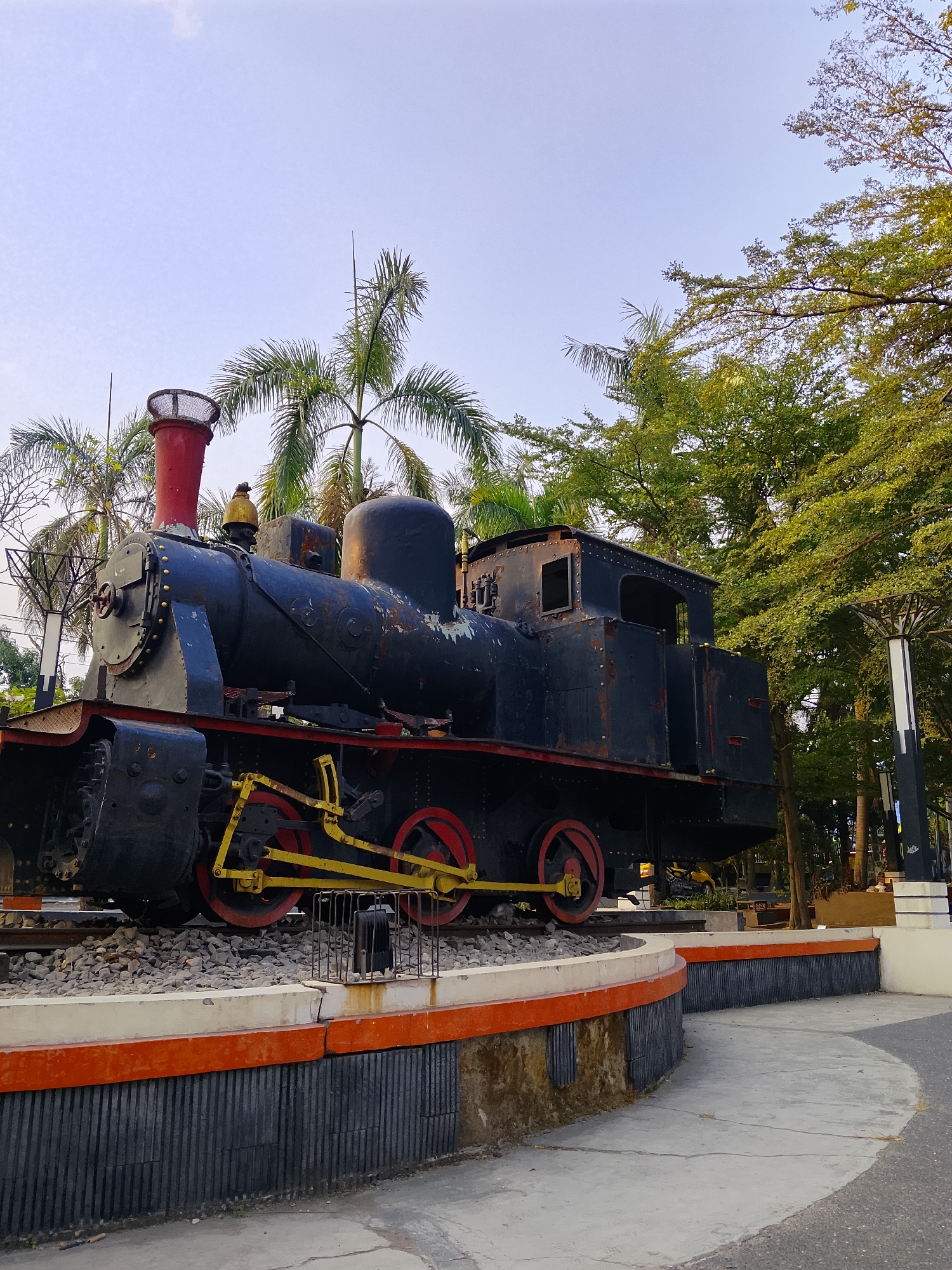 An old, black steam locomotive is displayed on a section of railroad tracks. The locomotive is parked on a circular, raised platform with a white and orange trim. The platform is surrounded by a paved walkway. Green trees, including palm trees, are visible in the background. The sky is clear and light blue. The image appears to be taken during the day, likely in the late afternoon given the lighting. There are no people or animals visible in the image. The locomotive is rusted in some areas, indicating its age. This locomotive is located in Balun, Indonesia.