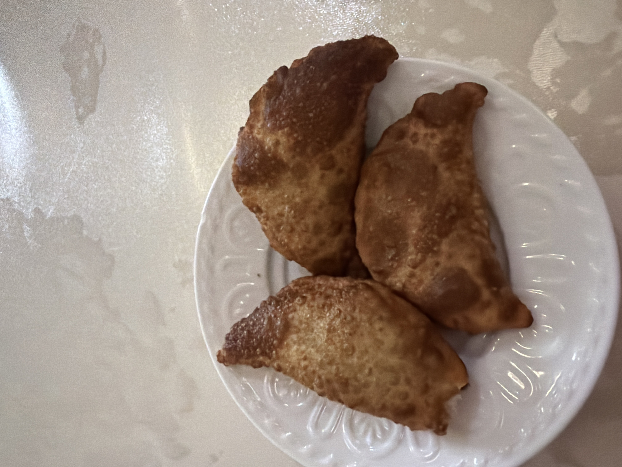 Three golden-brown fried pastries are arranged on a white, embossed plate. The pastries are crescent-shaped with crimped edges, suggesting they are empanadas or a similar filled pastry. They are positioned in a triangular formation on the plate. The plate itself rests on a textured, light-colored surface that appears to be a table with a subtle pattern. The lighting is bright, casting soft shadows. There are no people visible in the image. The image was taken in Malgobek, Russia.