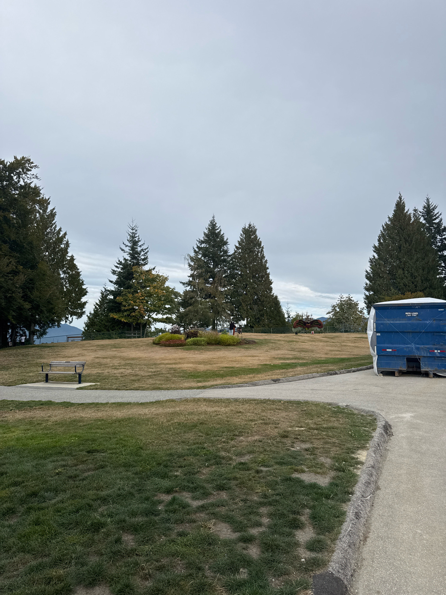 This image depicts a park scene under an overcast sky, likely in Burnaby, Canada, given the provided context. In the foreground, a concrete path curves around a patch of dry grass. To the left, a metal park bench with a wooden seat rests on a paved square. Further into the park, a slightly elevated grassy area is dotted with trees. In the middle ground, a decorative display, possibly a Halloween-themed spider made of plants and other materials, is visible. Several mature evergreen trees and some deciduous trees, some with autumn foliage, surround the park. On the right side of the image, a large blue roll-off dumpster with a white tarp covering its top is parked. In the background, rolling hills and distant mountains are faintly visible through the haze. A few individuals can be seen in the distance near the decorative display. The overall impression is a public park on a cool, cloudy day.