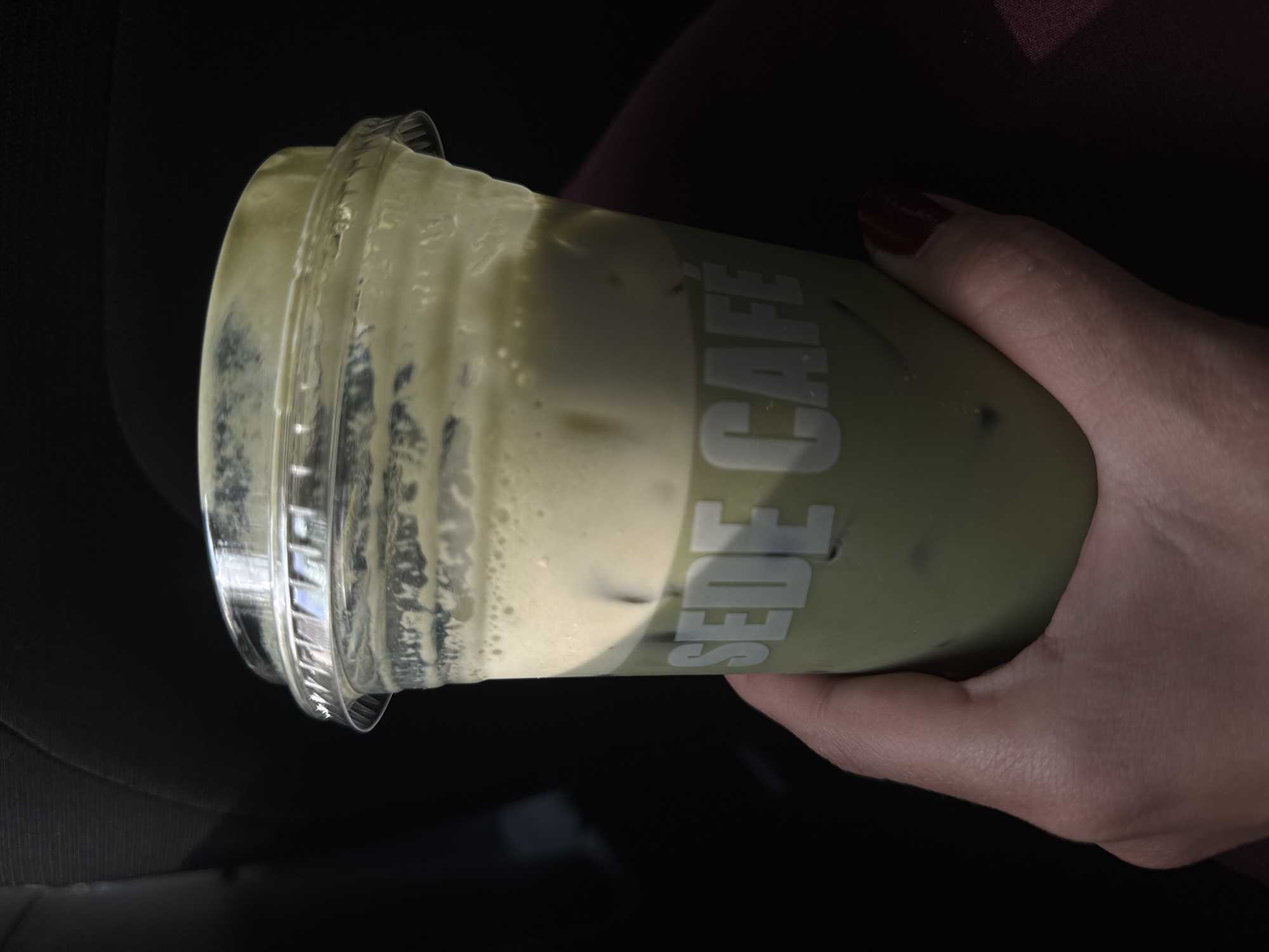 A person is holding a Sede Cafe iced matcha latte in their right hand. The hand has a red-painted nail. The drink is in a clear plastic cup with a clear lid. The drink appears to be matcha latte with ice. The backdrop shows a dark environment with hints of dark fabric/clothing. It is not possible to identify the location in Mexico City.