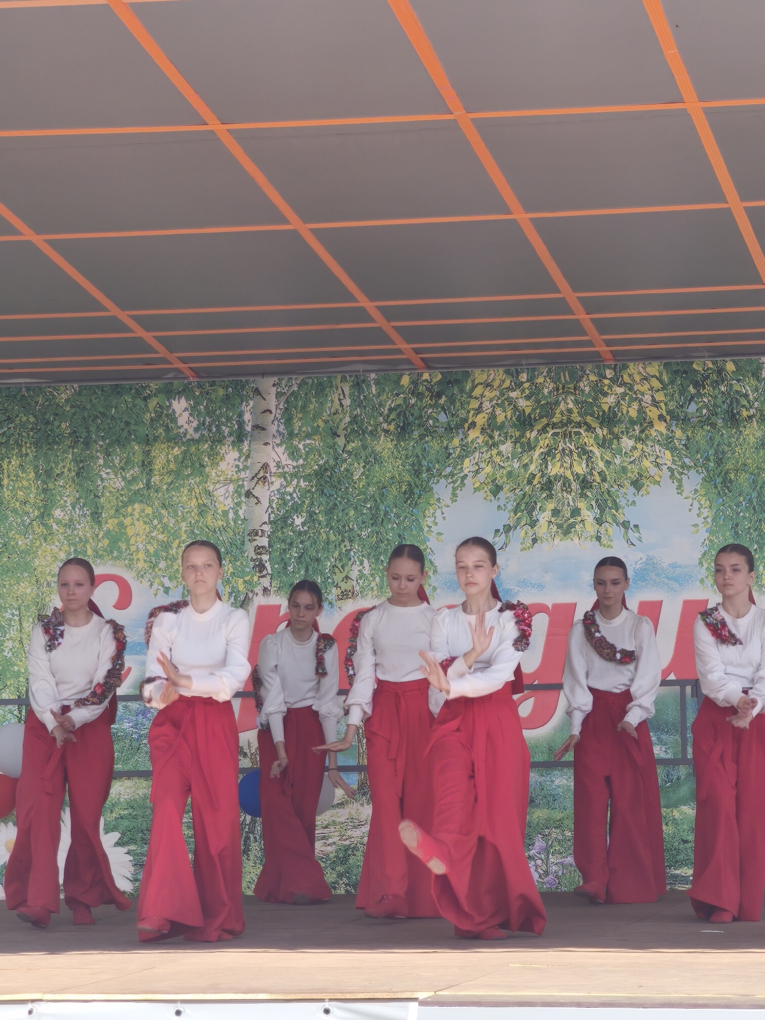 Eight young females, likely pre-teen or early teenagers, are performing a dance on an outdoor stage. They are dressed in matching costumes: white long-sleeved shirts with decorative trim around the neckline, and wide-legged red pants that flare out from the knee down. All of the dancers have their hair pulled back. The backdrop of the stage depicts a stylized forest scene with birch trees and green foliage, beneath a bright blue sky. In front of the dancers, large red letters are visible, though mostly obscured by the performers. Two balloons, one white and one blue, are partially visible behind the dancers on the left side of the stage. The stage itself appears to be made of wood, with visible mounting hardware on the front edge. Above the stage, an orange and gray striped awning provides shade. The event is taking place in the city of Kady, Russia.