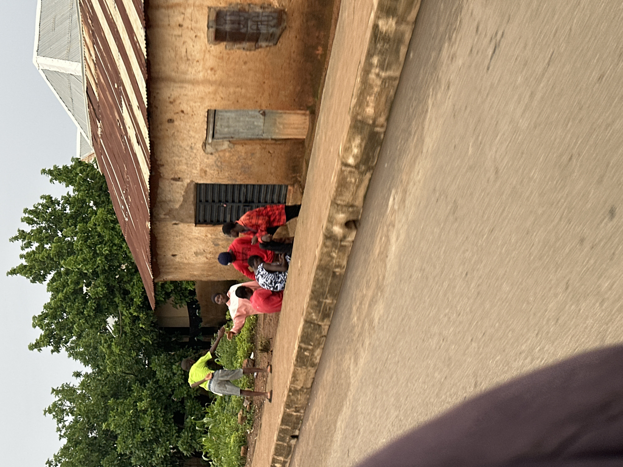 In Bukuru, Nigeria, a group of individuals is gathered by a dirt road. On the left, a person in a lime-green shirt and grey shorts stands, facing away from the camera with their right arm extended. To their right, a person in a pink and white top sits on the ground, facing the person in green and with their left arm extended. Behind them, a person in a red top is also seated. Further to the right, two more individuals are visible, one in a black and white patterned shirt and the other in a red and orange checkered shirt, both seated on the ground. Behind the seated individuals is a simple, mud-brick building with a corrugated metal roof. The building has a dark-colored door and a weathered wooden door on its facade, along with a barred window opening. The environment is characterized by lush green foliage bordering the road and the building. The road itself appears to be made of dirt or gravel, with a concrete curb running along its edge. The scene suggests a casual gathering or interaction on a typical day in the town.