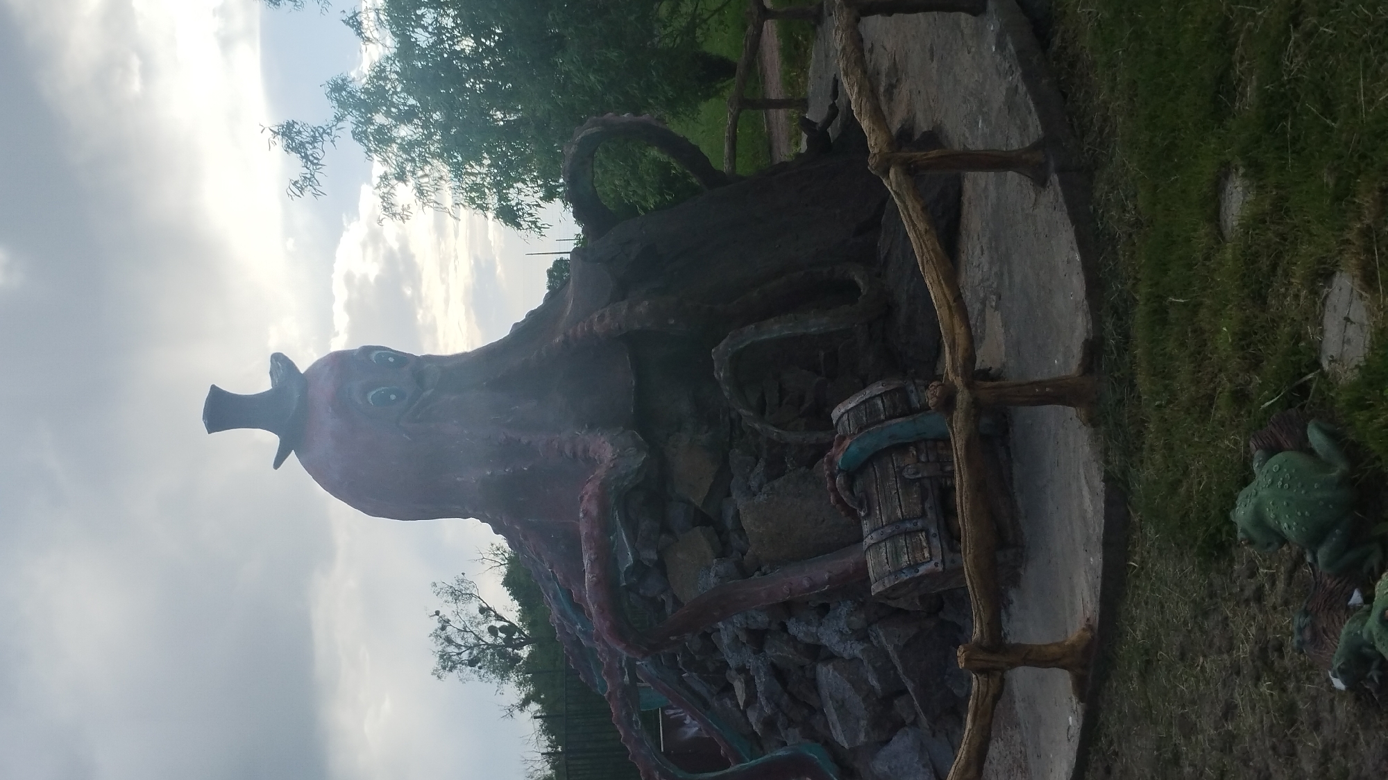 The image depicts a whimsical sculpture located in Khmilnyk, Ukraine. The sculpture features a large, cartoonish octopus wearing a top hat. It is positioned on a rock structure surrounded by a wooden frame. A wooden barrel and several frog figures are near the base. The background includes trees, grass, and a cloudy sky.