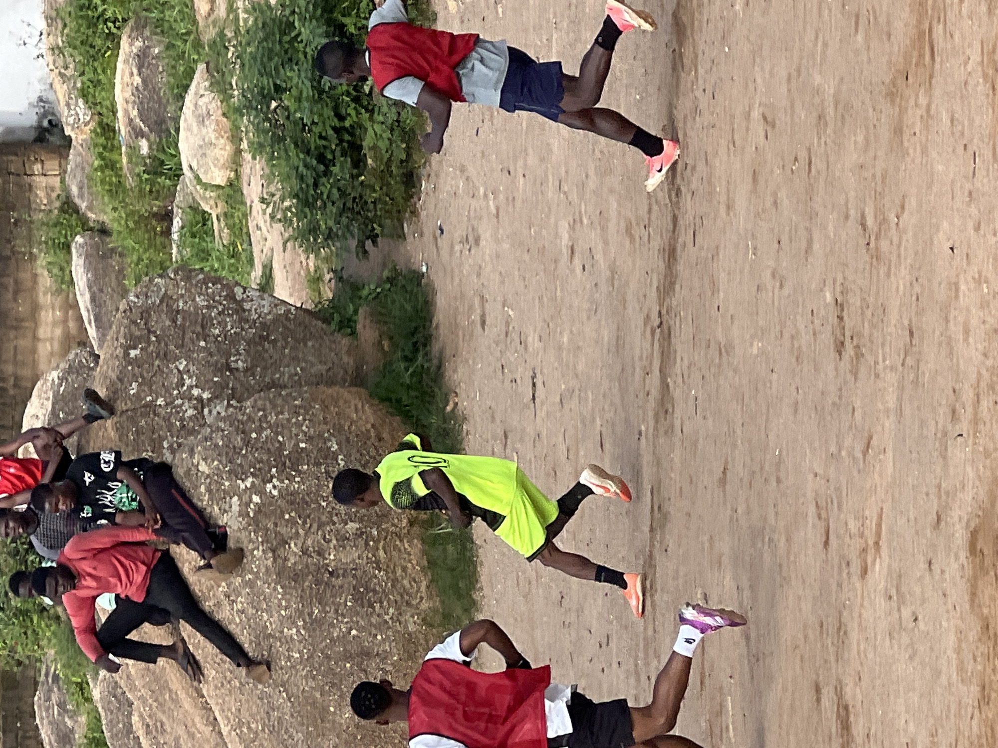 The image shows a group of young men in a casual setting. Two of them are sitting on a large rock, while three others are actively engaged in playing soccer on a dirt field. The environment appears to be outdoors, with rocks and vegetation visible in the background, suggesting a hilly or rocky terrain. The overall scene depicts a lively moment of recreation and youth activity.