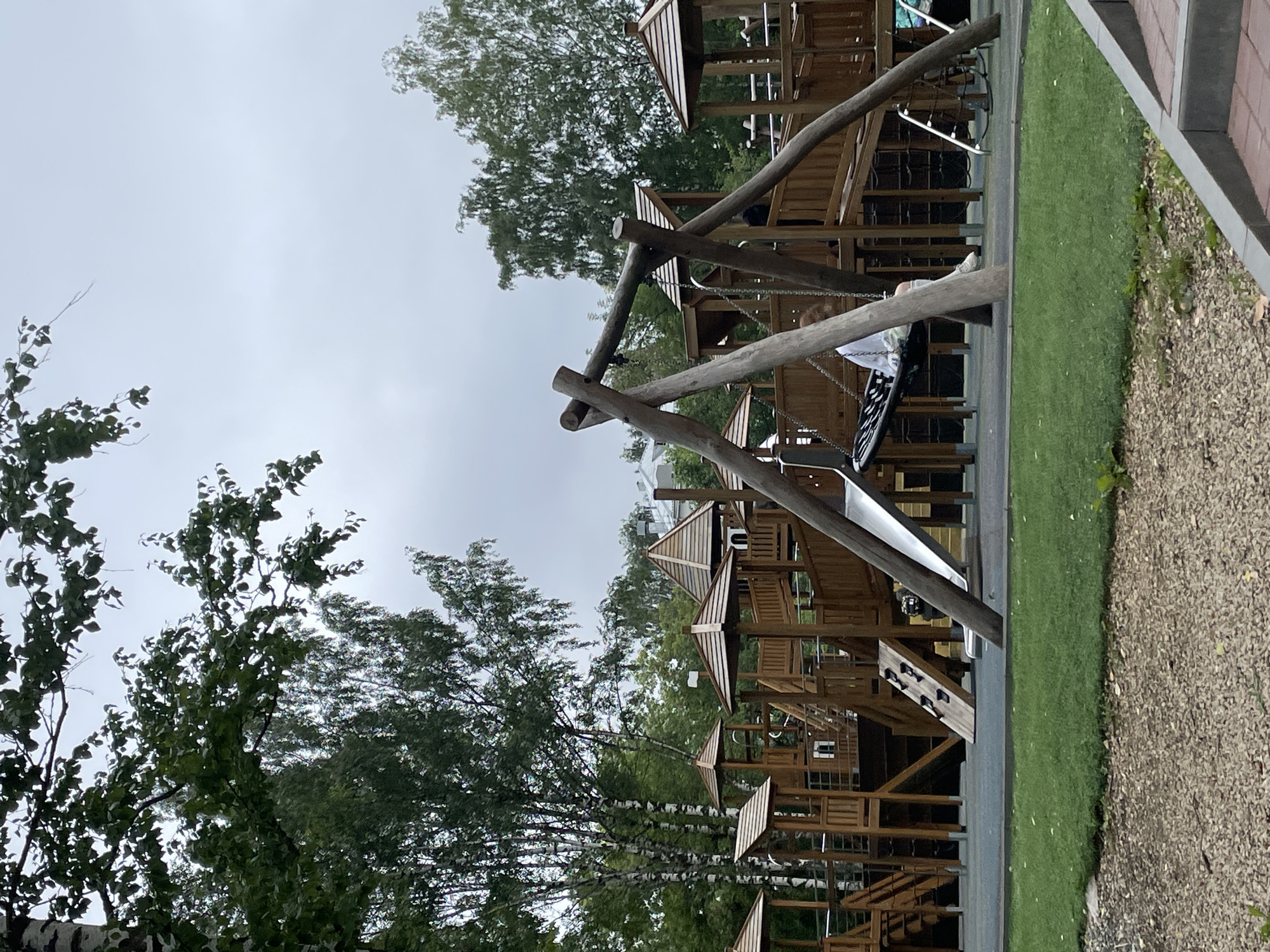 In Kirov, Russia, an outdoor wooden playground features multiple interconnected structures with slides, climbing walls, and swings. A child, wearing a white shirt and pants, sits on a black fabric swing. The ground is covered with soft, blue matting, transitioning to green grass and then to a surface of small stones and wood chips. Towering birch and other deciduous trees surround the playground, under an overcast sky. In the background, buildings are visible beyond the trees.