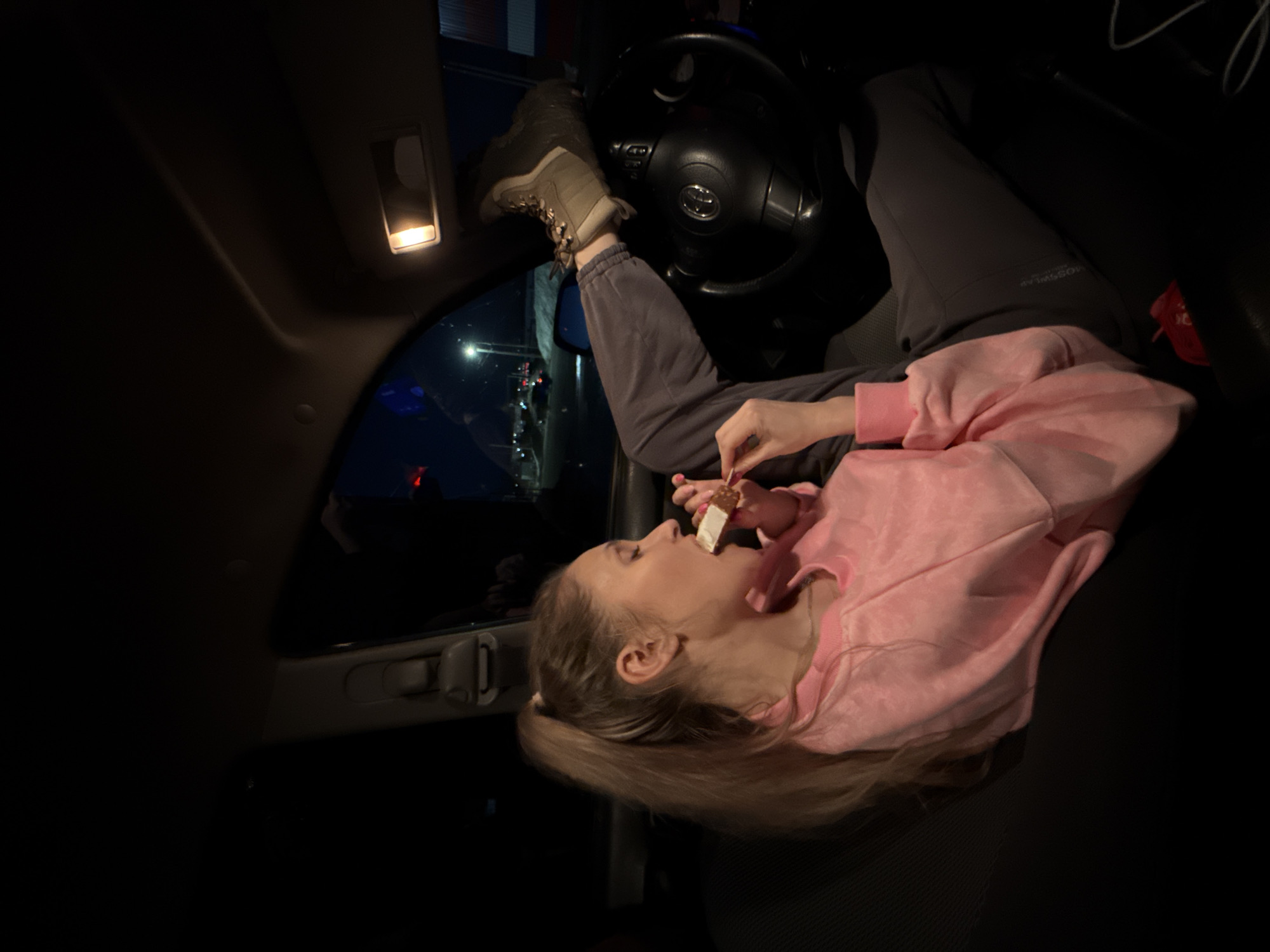 The image shows a young woman inside a car, possibly in a drive-through setting. She has her feet up on the dashboard or possibly the steering wheel, with one foot wearing a laced hiking boot. She is wearing gray pants and a pink sweatshirt. She is eating what appears to be an ice cream bar or similar snack held with a small wooden stick. The interior car light is on and there are external lights visible through the car window suggesting an urban setting. The steering wheel has the logo of Toyota on it. No evidence supports the location of Chaikovsky, Russia.