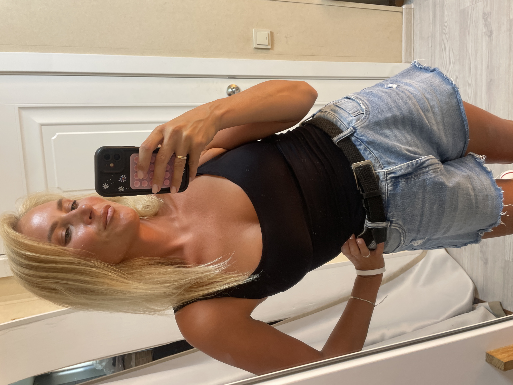 In Baranovka, Russia, a blonde woman is taking a mirror selfie in what appears to be a home environment. She is wearing a black tank top, denim shorts, a black belt, and a gold ring on her left hand. She is holding a black smartphone with a floral case and a pink pop socket. Her left arm is on her hip, and her right hand is holding the phone. The background shows a white door, a light-colored wall, and a mirror reflecting part of the room, including curtains.