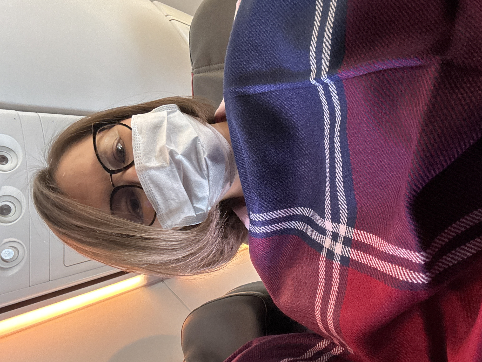 A woman wearing glasses and a white face mask is seated on an airplane. She is partially covered by a plaid blanket of red, blue and white. The interior of the plane can be seen in the background, including a control panel and seat. The location is not identifiable as Lyamtsino, Russia from the information given in the picture.