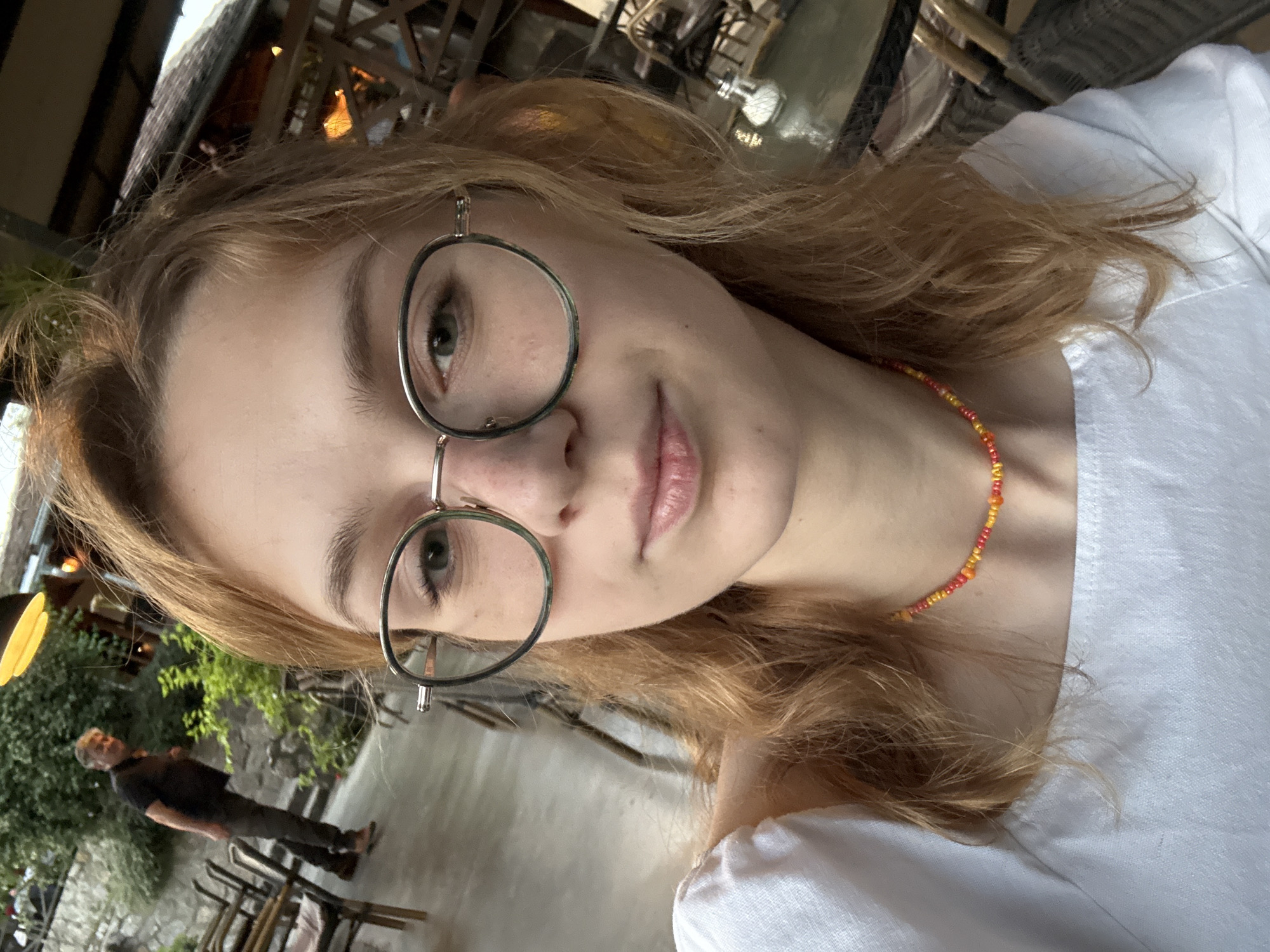 A young woman with light-colored hair is taking a selfie. She is wearing round glasses and a white shirt. A colorful beaded necklace adorns her neck. In the background, slightly blurred, is a man standing on a patio-like area with stone walls and greenery. There are chairs and tables visible in the background as well. The location appears to be an outdoor seating area, possibly in Kragujevac, Serbia. The general setting suggests a restaurant or cafe.