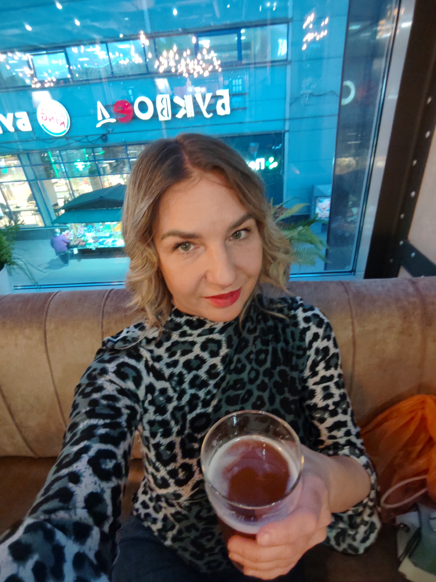 A woman is taking a selfie indoors, seated on a comfortable upholstered sofa or booth in what appears to be a restaurant, bar, or cafe. She has blonde, wavy hair, light-colored eyes, and is wearing red lipstick. Her attire is a long-sleeved top with a black and grey leopard print pattern. She holds a glass containing an amber-colored beverage, likely beer, with a foamy head.
The setting is characterized by a large window behind her, offering a view of an urban environment, suggesting it's evening or night due to the reflections of indoor lighting and brightly lit signs from outside. The scene outside appears to be a commercial area, possibly a shopping mall or a street with multiple establishments. An orange mesh bag or fabric is visible to her right.
Visible text on the window, reflected from outside signs, includes a reversed "Burger King" logo (specifically "КИнг") and other reversed Russian letters. A prominent reversed sign displays "ДЯБОРЯА", which, when correctly reversed, reads "АЯРОБАД", likely a brand name. Partial reversed letters "ВА" and a character resembling "С" are also visible.
The woman looks directly at the camera with a neutral to slightly pleasant expression. The atmosphere suggests a casual social outing in Saint Petersburg, Russia.