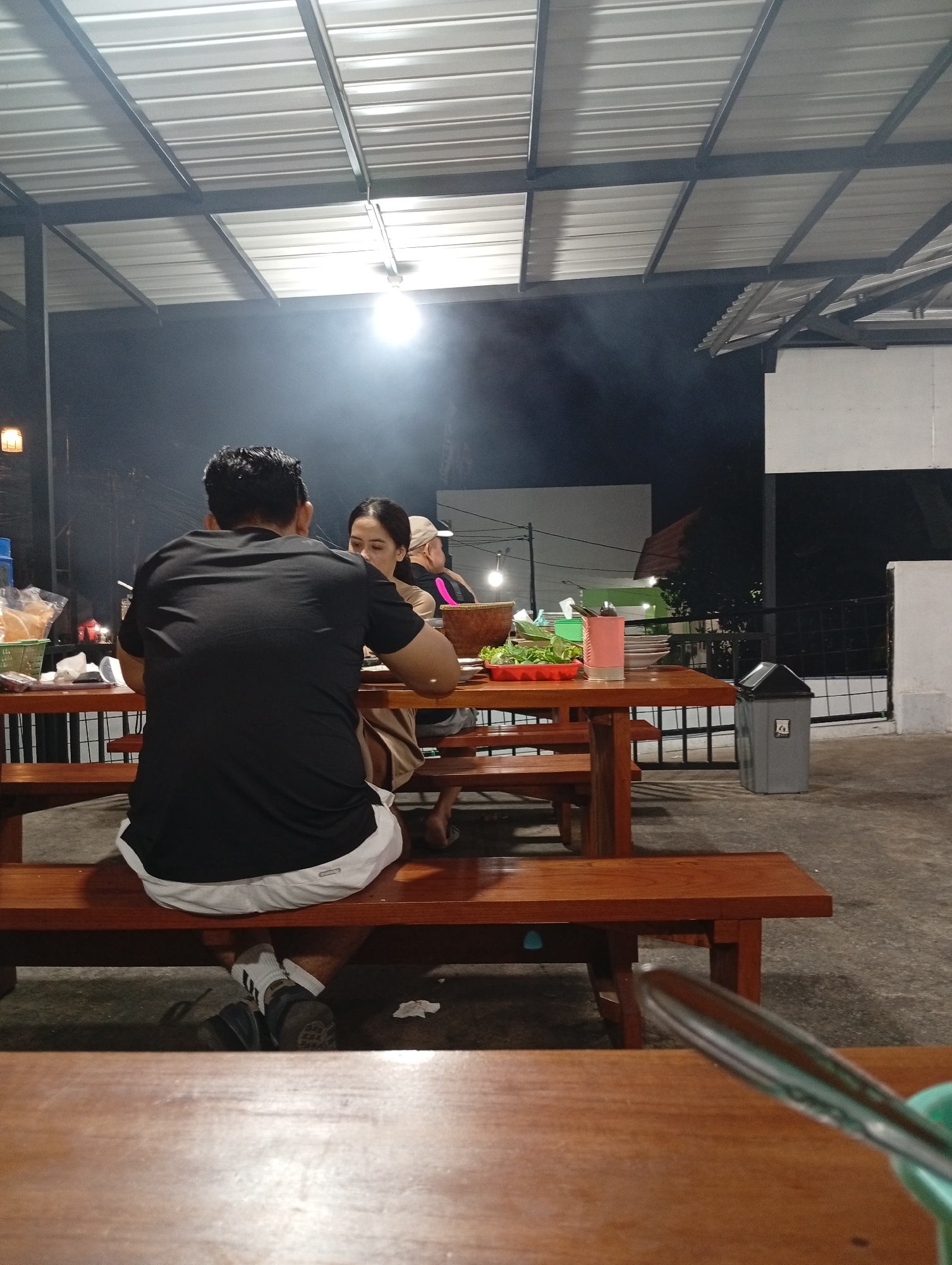 The image depicts an outdoor dining area at night, likely a restaurant or warung, in Pacet, Indonesia. A man and a woman are seated at a wooden picnic table, facing away from the camera, engaged in a meal. The man is wearing a black t-shirt and white shorts, while the woman is wearing a light-colored top. A variety of food items are spread across the table, including a bowl of vegetables, plates, and a pink container. In the background, a white building and a black fence are visible, along with a trash can. The scene is illuminated by overhead lights, creating a smoky atmosphere.