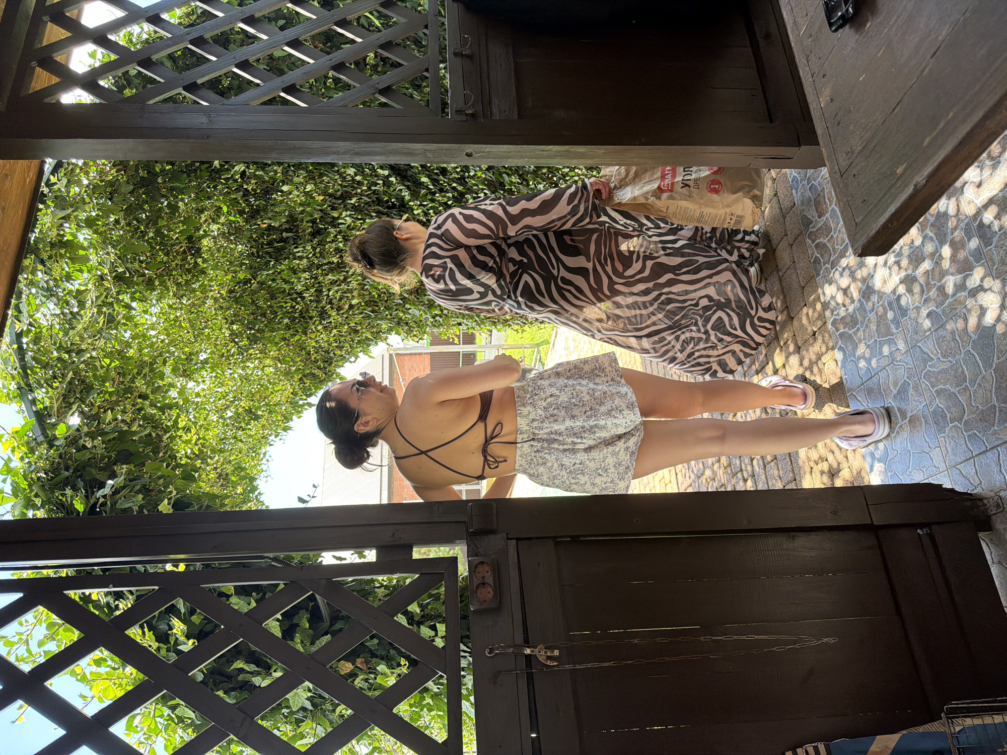 This media file shows two individuals outdoors, likely in a backyard or garden setting, under a wooden pergola or gazebo. The scene is lush with green foliage, suggesting a warm weather or summer day.
In the foreground, a person is seen from the waist down, wearing a light-colored floral skirt and white sandals. They are standing on a patterned tiled surface that transitions to cobblestone.
Behind them, another individual is visible from the shoulders up, wearing a sheer, long-sleeved dress with a zebra print. This person appears to be interacting with an object behind them, which is partially obscured but looks like a large bag, possibly containing charcoal or briquettes, as suggested by visible text on the bag that reads "УГОРЬ ДРЕВЕСНЫЙ" (Ugor' Drevesnyy - Wooden Eel) and other Cyrillic characters.
The overall impression is a casual, outdoor gathering or preparation for an activity like grilling. The lighting suggests it is daytime, possibly late afternoon given the angled sunlight creating shadows. The atmosphere appears relaxed. The wooden structures, abundant greenery, and the tiled and cobblestone ground provide context for the outdoor location.