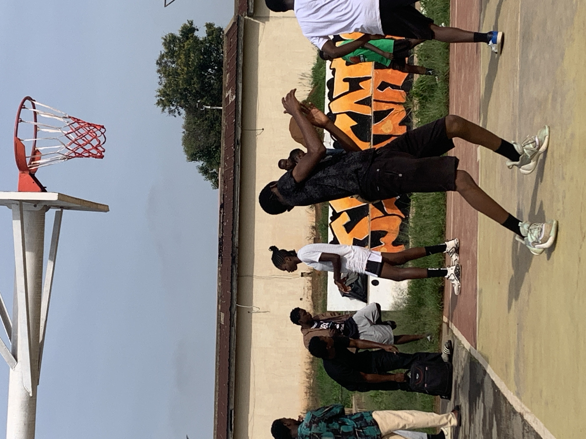 The image depicts a group of young people on an outdoor basketball court in Gura Top, Nigeria. The court has a reddish-brown and yellow surface with white lines. A basketball hoop with a red rim and white net is visible on the left side of the frame.
Several individuals are scattered across the court, with some appearing to be engaged in a game or practice. One person in the foreground, wearing a black t-shirt and black shorts, is captured mid-motion, possibly playing basketball. Other individuals are standing or sitting, some observing.
A building with a textured, light-colored wall and a rusted metal roofline forms the backdrop. Behind the building, lush green trees suggest an outdoor, possibly warm-weather setting. The sky is a clear, pale blue, indicating daylight, likely late morning or afternoon. The overall mood appears casual and active, with no overt displays of strong emotion.
Some of the individuals are wearing athletic attire. One person is wearing a white t-shirt and black shorts, while another is in a green shirt. Graffiti featuring orange and black designs is visible on a section of the wall in the background, adding a touch of urban art to the scene. There is no discernible text visible in the image.