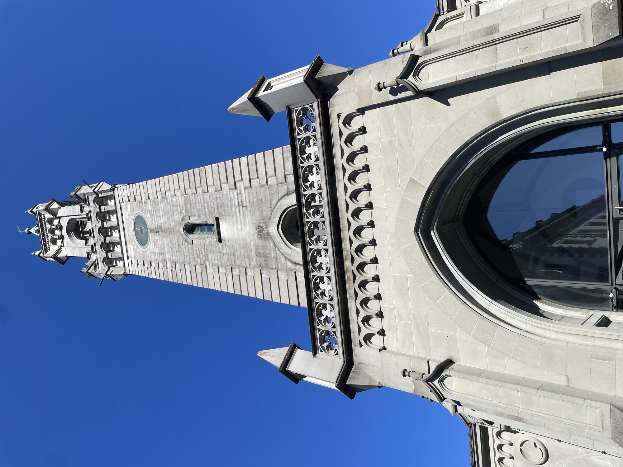 A low-angle shot captures the imposing stone clock tower of the Konstanzer Münster, or Constance Minster, in Constance, Germany. The tower, constructed from light gray stone blocks, rises dramatically against a clear, vibrant blue sky. Intricate architectural details are visible, including a decorative balustrade with circular patterns, arched window openings, and sharp, pointed spires atop smaller turrets. A round clock face with a green tint is centrally positioned on the tower. The lower section of the tower features a large, arched window with a dark frame, reflecting the surrounding architecture. The overall impression is one of historical grandeur and architectural significance.