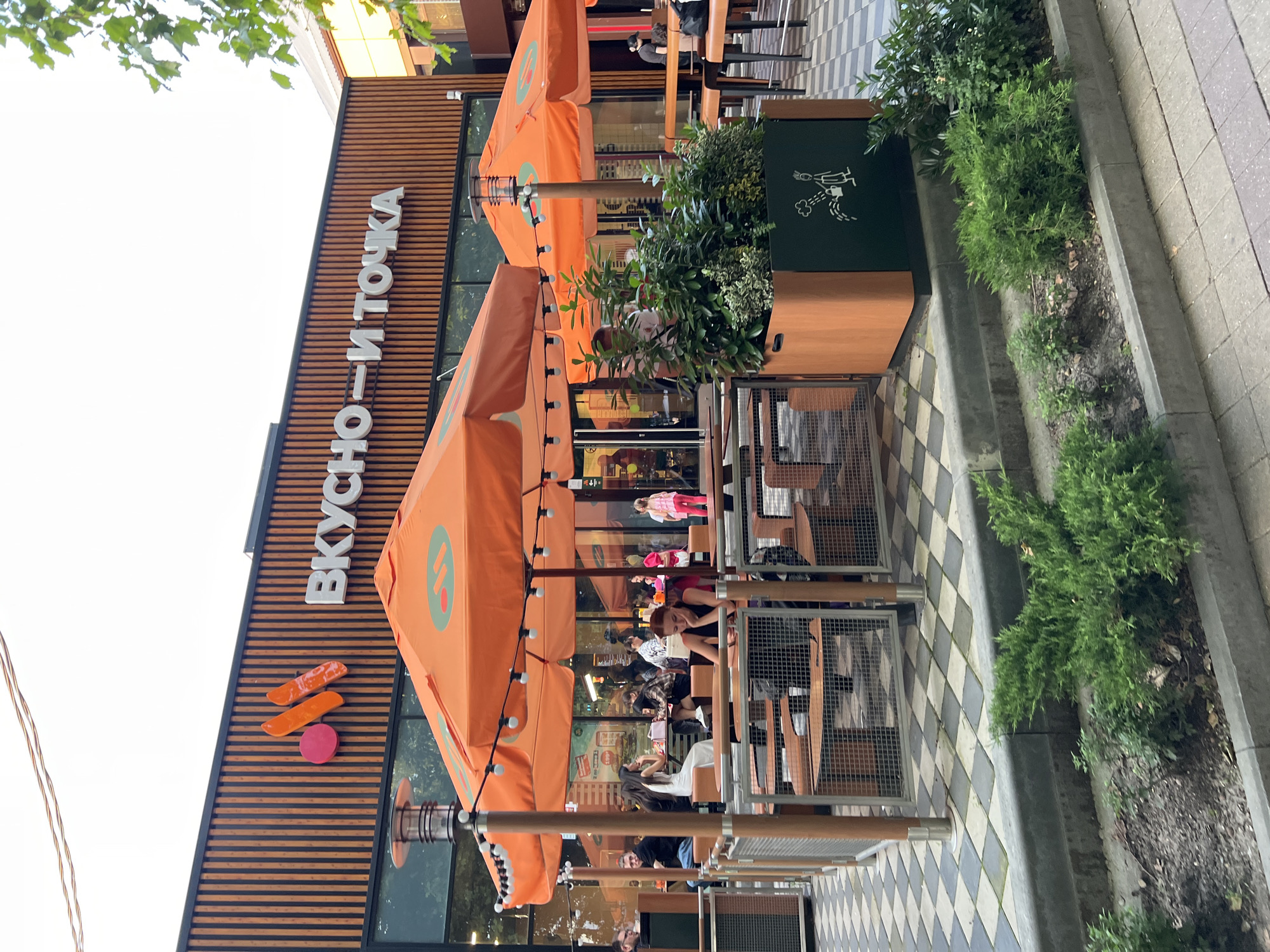 This image shows the exterior of a "Vkusno i tochka" restaurant in Krasnodar, Russia. The restaurant has a modern design with vertical wooden slats on the facade and a prominent "Vkusno i tochka" logo. There are orange umbrellas providing shade over outdoor seating areas, furnished with wooden tables and chairs. Several people are seated at the tables, some interacting and others looking at their phones. A woman in a black top is visible in the foreground, looking down at her phone. In the background, more patrons are seated, and some are queuing inside the restaurant, which has large glass windows. The ground is paved with a checkered pattern of light and dark tiles. A planter box with greenery is situated in front of the restaurant. The overall scene depicts a casual dining environment on an overcast day, with clear evidence of an active establishment.