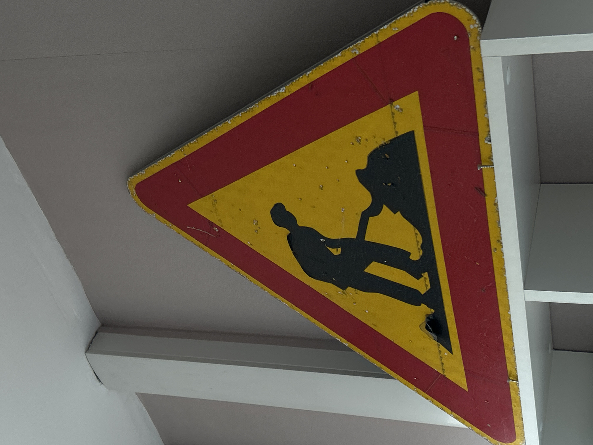 An old, triangular road sign hangs from a ceiling. The sign has a yellow background, a red border, and a black silhouette of a man working in a mine. The sign appears to be in a state of disrepair with rust and wear visible. The environment includes a white ceiling and architectural elements suggesting an indoor location. The city and country are not discernible from the image.