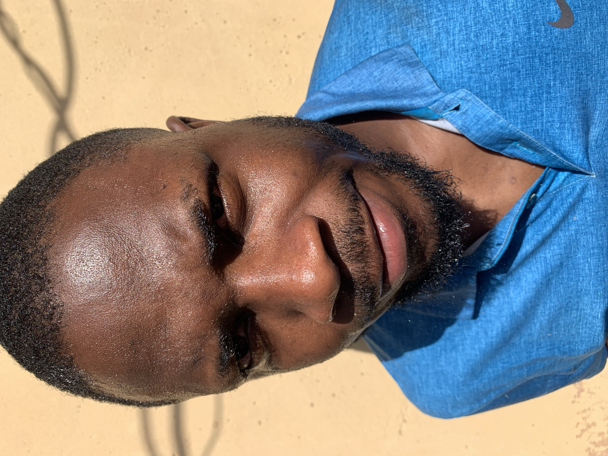 A close-up, angled portrait shows a Black man with closed eyes, a short haircut, and a beard. He is wearing a light blue, button-down shirt, possibly a polo shirt. The background is a light tan or beige wall with shadows cast on it. The lighting is bright and appears to be natural sunlight. There are no readily identifiable elements to determine the location in the city of Rafin Par, Nigeria.