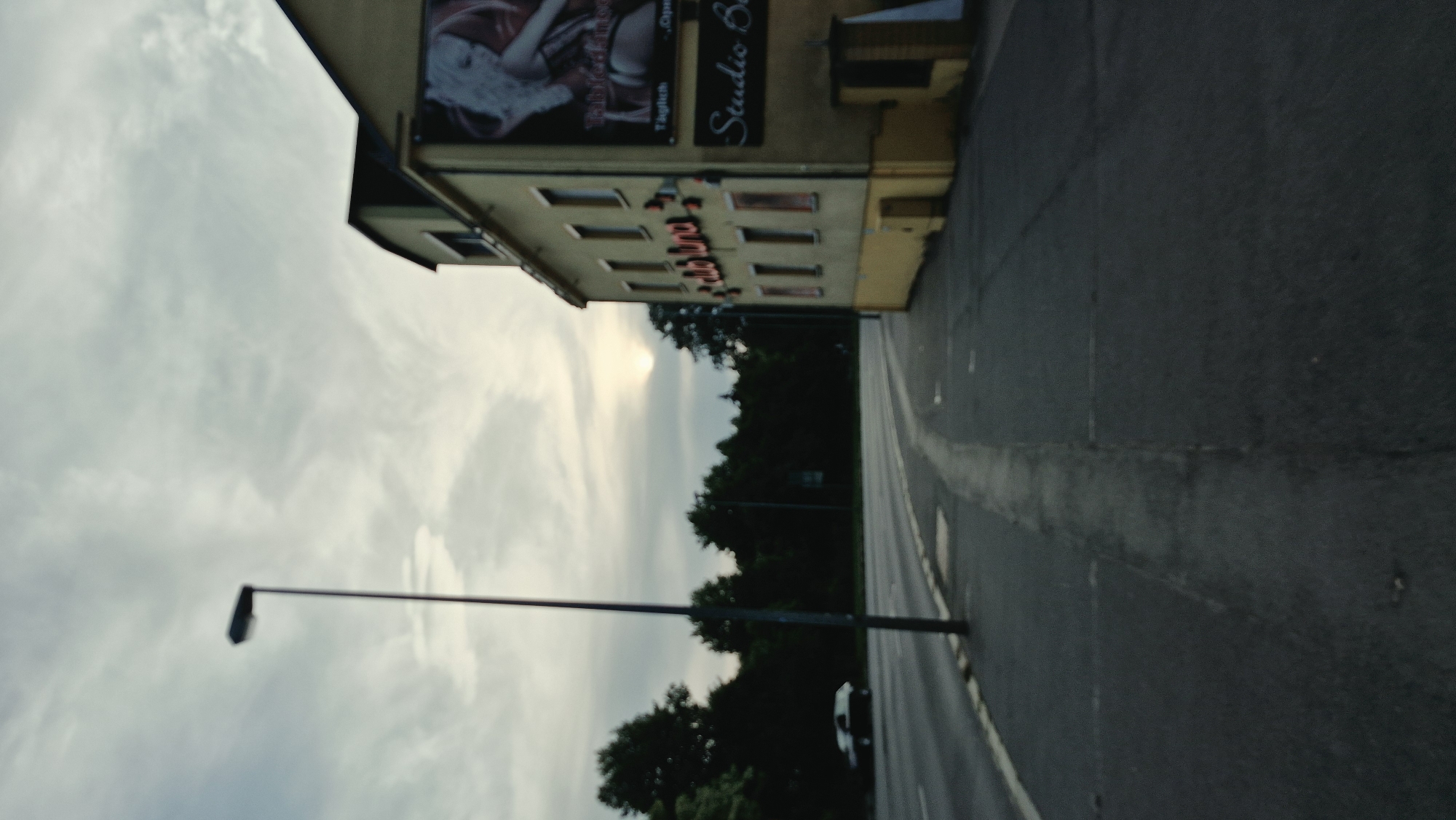 The image shows a street scene in Chemnitz, Germany. A light-colored building with yellow trim stands on the right side of the frame, featuring a large banner with an image of a blonde woman and text that reads "Tabfofdinsn Täglich". Below this, another sign reads "Studio B". Red neon lights spelling out "club luna" are visible on the building's facade. A street light stands on a pole in the foreground, and a road curves to the left, with a car parked on the left side of the road. A line of dark trees forms a backdrop, with the sun partially obscured by clouds in the sky. The scene appears to be a view of a street with a club and a studio.