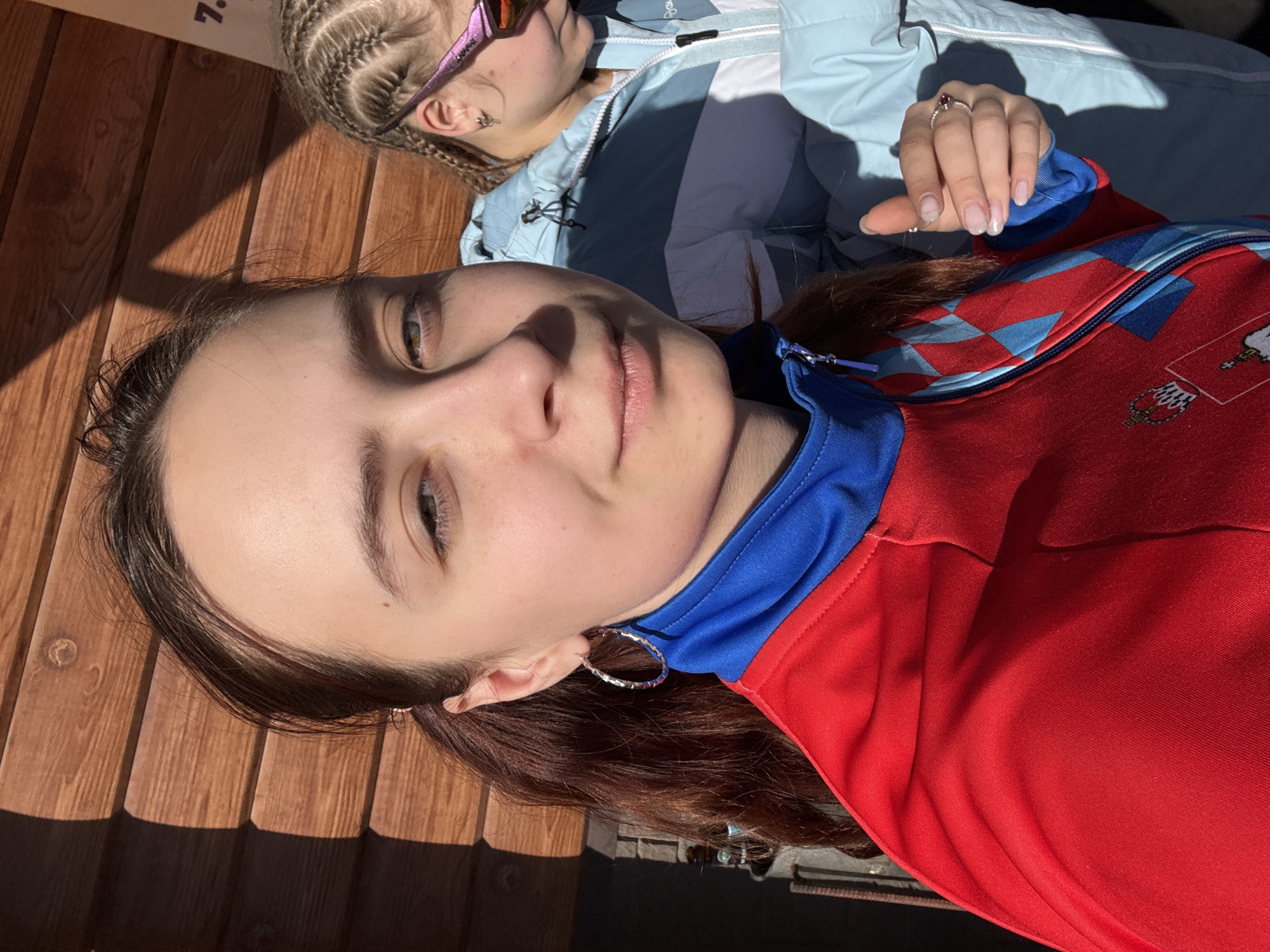 The image shows two young women from a low-angle view. One is wearing a red jacket with a blue collar and a pattern on the shoulder, while the other wears a light blue jacket. The first woman has brown hair and is wearing hoop earrings. The second woman has braided blonde hair and is wearing sunglasses. The background appears to be a wooden structure. A partial sign in the top-left corner suggests this might be a public place or venue. Based on the clothing and apparent location details, it can be surmised that the picture was taken in Gory, Russia.