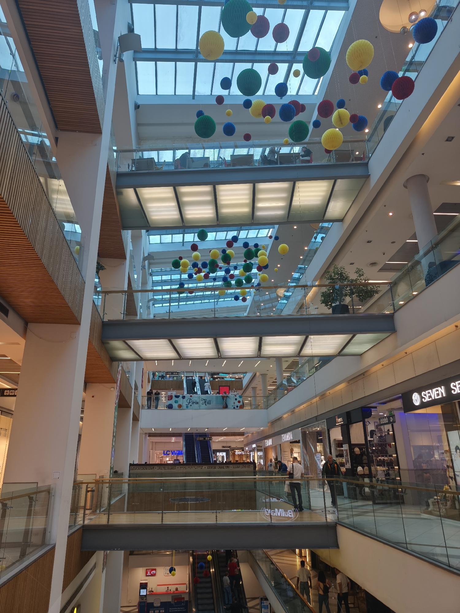 The image shows the interior of a multi-level shopping mall in Varna, Bulgaria. The architecture includes glass railings, white pillars, and a multi-tiered atrium topped by a skylight. Above, colorful spherical decorations are suspended from the ceiling. People are seen browsing and walking along the various levels of the mall, with storefronts like "Seven Seconds" and "Bershka" visible. Escalators connect the floors, and signage like "Easy Pay" and "LC Waikiki" is present. Text in Bulgarian is also visible.