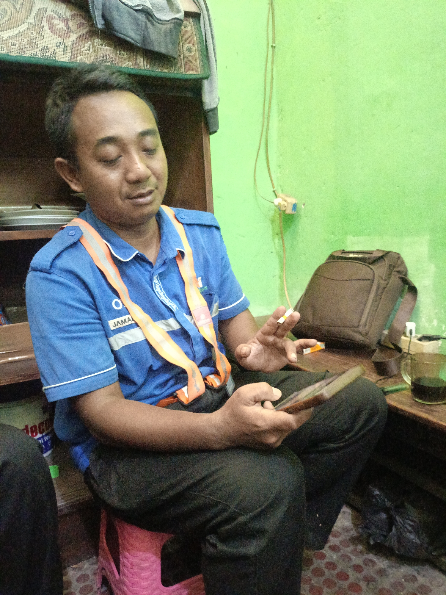 A man, identified as Jamal, sits on a pink plastic stool in an indoor setting. He is wearing a blue shirt with an orange safety vest over it. His right hand holds a cigarette, and his left hand is holding a tablet computer. A brown bag is visible behind him, and a glass of coffee is on a wooden table to his right. The wall behind him is painted green. The scene appears to be in Kidul Dalem, Indonesia.