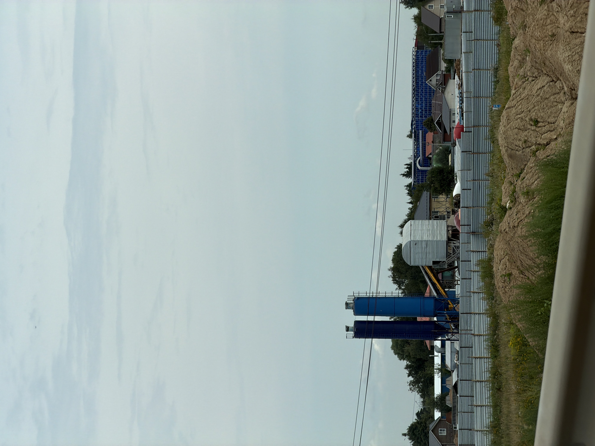 The image depicts an industrial area on the outskirts of Durykino, Russia. In the foreground, a mound of sand and dry grass are visible. A corrugated metal fence runs horizontally across the midground, separating the foreground from the industrial structures. Behind the fence, two tall, cylindrical blue silos stand prominently, with a smaller, grey, hopper-like structure next to them. A conveyor belt extends from the silos. Several houses, some with red roofs, are scattered behind the industrial equipment. A large, modern building with a blue glass facade is visible in the background, suggesting a mixed-use or commercial area. Power lines are strung across the sky, and the sky itself is overcast with scattered clouds. A tiny, dark object, possibly an airplane, is visible in the upper left quadrant of the sky. The scene appears to be a construction or industrial zone under a cloudy sky.