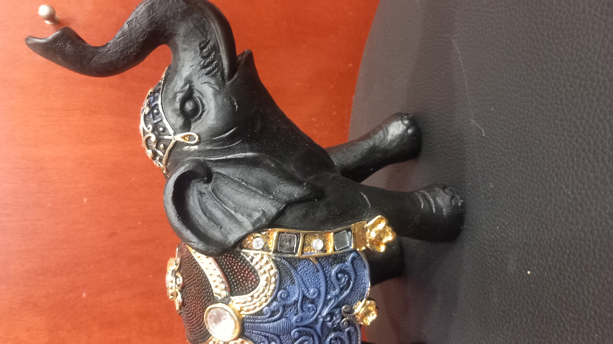 A close-up, eye-level shot shows a black, decorative elephant figurine with ornate gold and blue saddlecloth. The elephant has its trunk raised. It is positioned on a dark, textured surface, possibly leather. The background is a solid, warm orange-red color. The image is undated and its location in the city of Kos, Greece, is not specified.