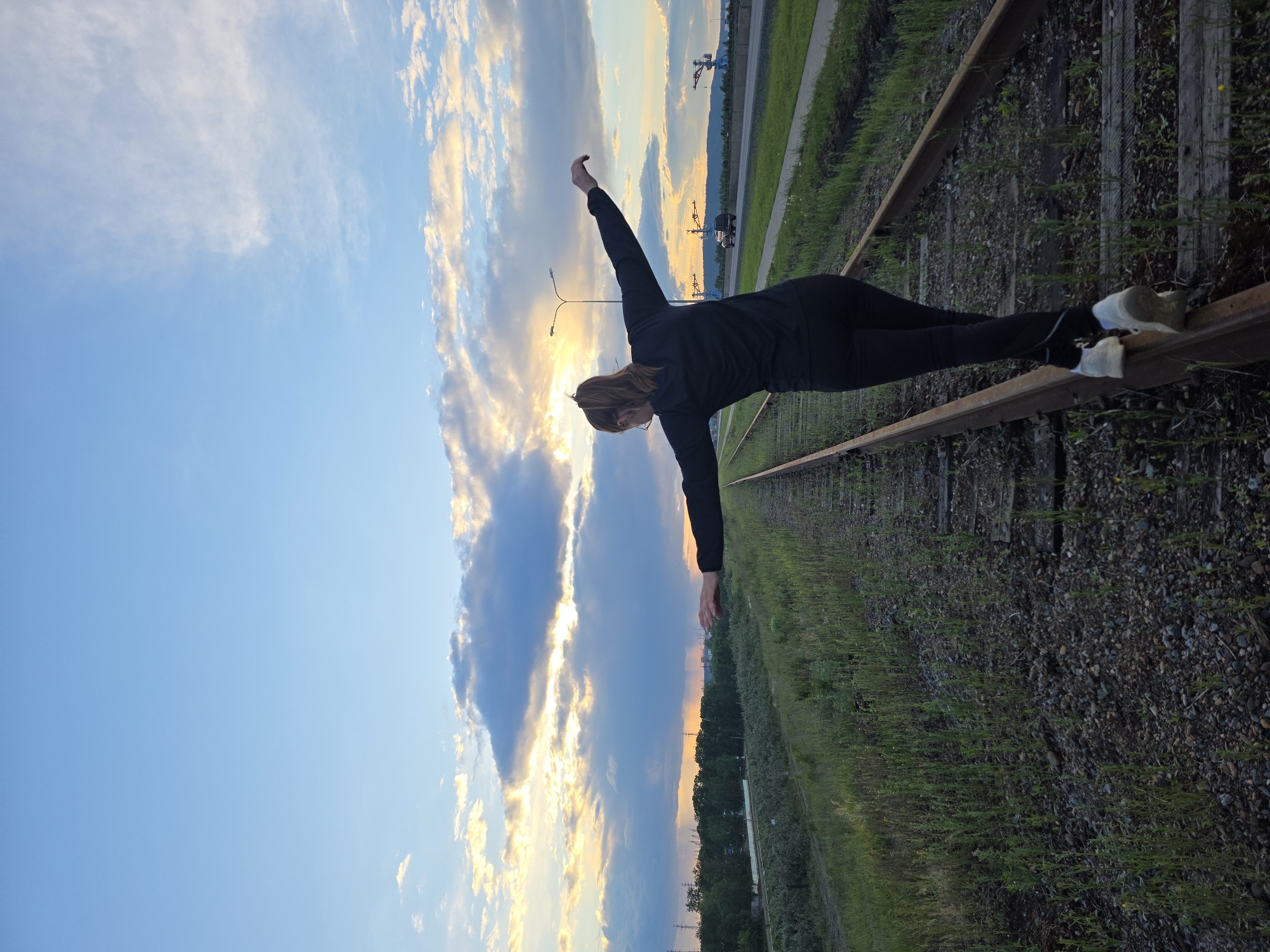 A woman is performing a balancing yoga pose on a set of train tracks. She is wearing a black top and leggings, and sneakers. Her body is parallel to the train track, with one foot and one hand resting on the rail. Her other arm is raised in the air. The background is a cloudy sky at sunset with a crane visible. The tracks are surrounded by gravel and vegetation. The picture was taken in Chaikovsky, Russia.