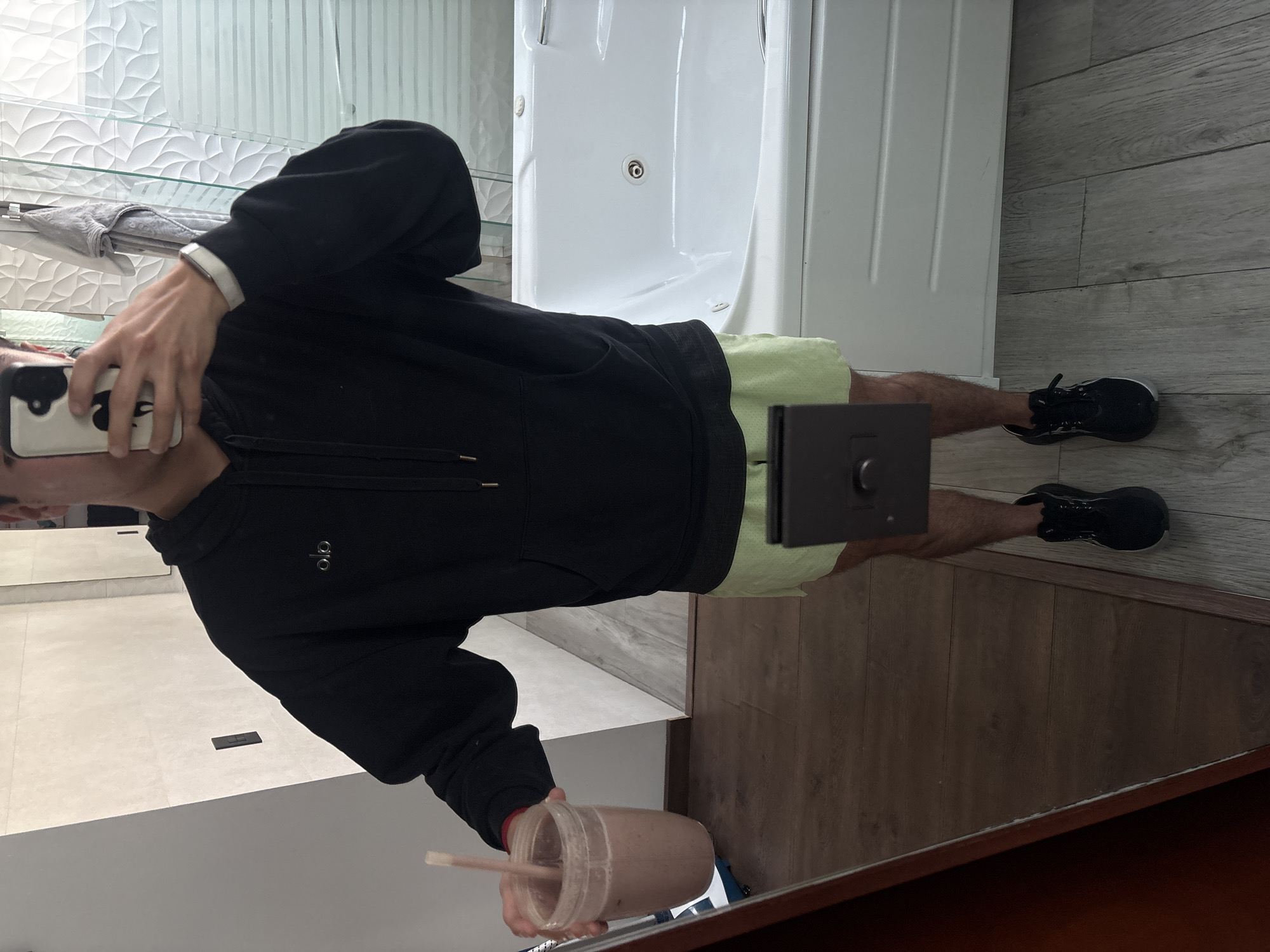 A person takes a mirror selfie in a modern bathroom located in San Bernardino Tlaxcalancingo, Mexico. The individual, dressed in a black hooded sweatshirt (hoodie) with a small, indistinct logo on the chest (possibly "Alo"), light green shorts, and black athletic shoes, holds a white smartphone with a distinctive black and white cow-print case in their left hand, framing the shot. A silver-colored band, possibly a smartwatch, is visible on their left wrist. Their right hand holds a clear plastic cup containing a brown liquid, likely a smoothie or protein shake, with a straw.
The background reveals a contemporary interior featuring a white wave-textured tiled wall behind glass shelves, one of which holds a grey folded garment. To the right, a white bathtub or shower unit with a visible drain stands against the wall. The flooring transitions from clean white tiles, where the person is standing, to wood-look laminate tiles further back. A small black rectangular object rests on the white floor tiles between the person's feet. The overall lighting is bright and even, indicating an indoor setting. No specific time of day or weather is discernible from the image, and the person's face is mostly obscured by the phone, showing a neutral expression focused on taking the photo.