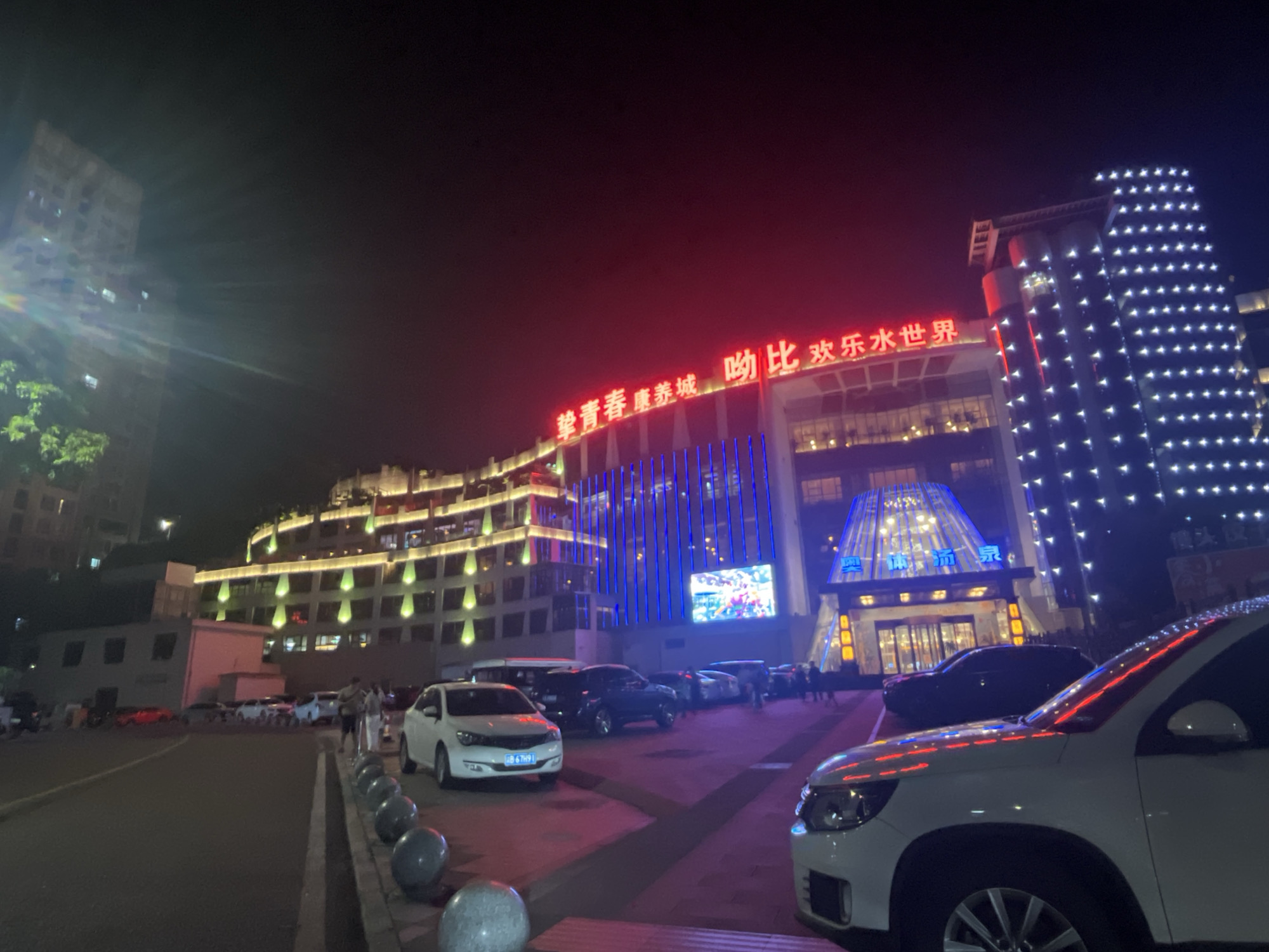 This nighttime image depicts the brightly illuminated "Olympic Sports Hot Spring Health City" (奥体·泓泉健康城) in Jiulongpo District, Chongqing, China. The scene focuses on a multi-story commercial complex with a parking lot in the foreground.
The central building features a prominent red neon sign displaying Chinese characters that translate to "Youthful Health City Kiss-me Happy Water World" (挚青春 康养城 吻比 欢乐水世界). Below this sign, vertical blue LED lights adorn the building's facade. To the right, another section of the complex is covered with a grid pattern of white, dot-like lights. The main entrance, visible on the far right, is a glass dome structure with additional Chinese characters "奥体•泓泉" (Olympic Sports Hot Spring) above it, illuminated in blue. To the left of the main complex, a separate multi-story building is lit with yellowish-green lights along its balconies and upper floors. In the far background on the left, a tall, less illuminated residential-style building is partially visible.
A paved parking area occupies the foreground and midground, populated by several vehicles. A white SUV, possibly a Volkswagen Tiguan, is parked on the far right, with its front end partially cut off by the frame. Another white sedan with a Chongqing license plate (渝B 67H9) is parked on the left side of the lot. Numerous other cars, predominantly dark-colored sedans and SUVs, are parked throughout the lot. Several scooters or motorcycles are also visible near the base of the building on the left.
Multiple individuals are observed walking in the parking area and on the sidewalks near the complex's entrance. The ground features grey spherical bollards lining the edge of the parking lot, separating it from the roadway. The sky is dark, indicating the time of night, and light flares are present in the upper left portion of the image, likely from ambient light sources interacting with the camera lens.