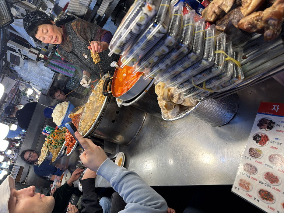 This image captures a bustling, brightly lit food stall within a traditional market in Seoul, South Korea.
In the foreground, a stainless steel counter extends across the scene. To the bottom left, a menu with Korean text and illustrated dishes, including prices, is visible. A person in a light blue long-sleeved shirt holds a black smartphone horizontally, appearing to photograph or record the scene. Partially visible to the far left is a male in a white bucket hat, seen in profile, looking towards the right.
At the center-right, an elderly Asian woman, likely the vendor, stands behind the counter. She wears a patterned head covering and a grey, patterned top with a dark apron. Her left hand, gloved, holds a skewer with multiple pieces of eomuk (fish cake), while her right hand uses tongs to retrieve an item from a large, shallow metal pot of simmering eomuk skewers. Adjacent to this, a deeper metal pot contains bright red tteokbokki (spicy rice cakes).
Stacked prominently in the foreground, next to the vendor, are numerous clear plastic containers of kimbap (Korean rice rolls), secured with yellow rubber bands. Next to these stacks, a silver-colored container holds steamed dumplings (mandoo). A tray of dark, cooked meat items is also visible to the right of the kimbap.
Further back, three customers are seated at the counter. On the left, a person with glasses and dark hair is visible behind multiple plates of Korean dishes, including kimchi, japchae, and a large bowl of white noodles. A green soda can rests among the dishes. Another customer is partially visible to the right of the first seated customer. The background features more market elements, including additional stalls, overhead white bowl-shaped light fixtures, and glimpses of other individuals, indicating a busy atmosphere.