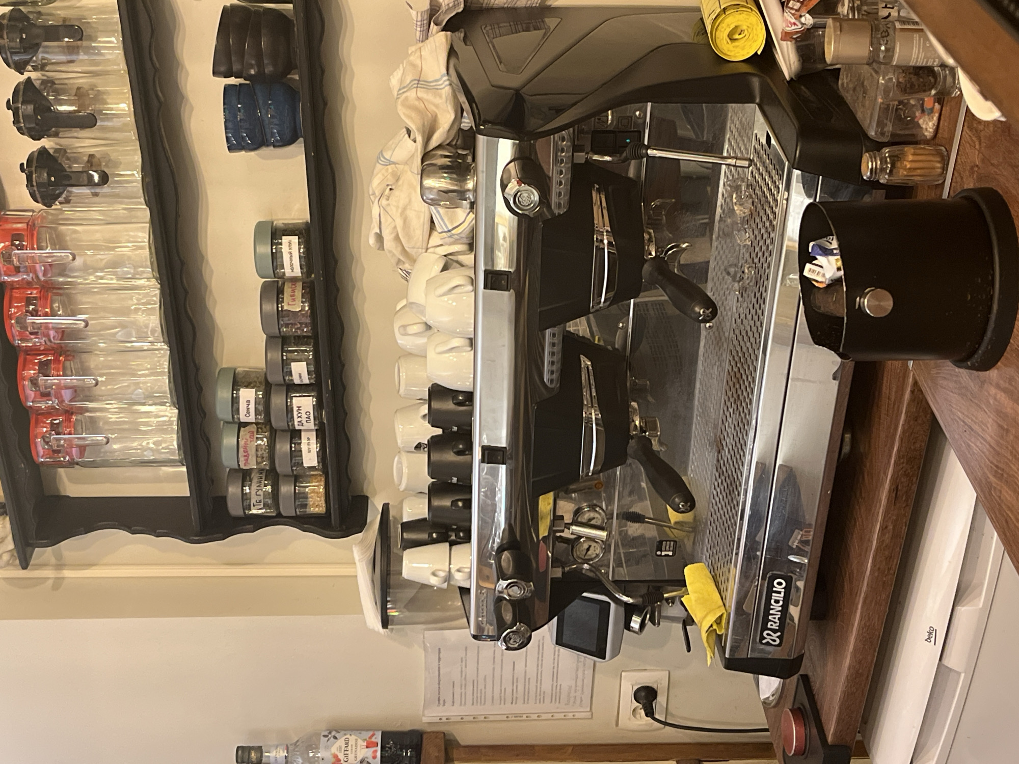 The image shows a commercial coffee machine, likely in a cafe or restaurant. It is a Rancilio brand machine and is positioned on a wooden counter next to a white refrigerator, possibly a Beko brand. Adjacent to the coffee machine is a collection of small, labeled spice jars organized on a wall-mounted shelf. Further to the left, there are several clear containers with black and red lids. Above the coffee machine are stacked white and black coffee cups. A black trash receptacle is placed beside the machine. A bottle of Giffard syrup is at the bottom left. The photograph's angle and composition suggest a tight, functional space within a food or beverage establishment, and based on the text that is read, the spices jars have "Сенча" and "ДА ХУН ПАО" written on them, which can mean Budva, Montenegro, or another Eastern European country.