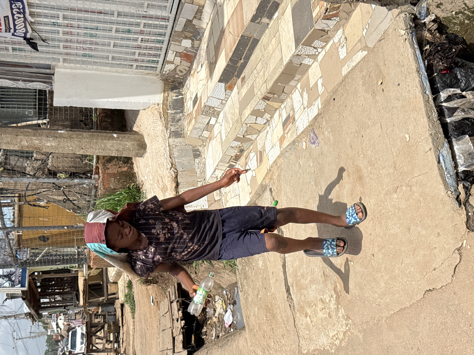 A young person stands on a concrete surface next to a gutter. The person wears dark shorts, a patterned shirt, and sandals, and holds a plastic water bottle. The background contains urban elements, including buildings and trash. The location appears to be in Abuja, Nigeria.