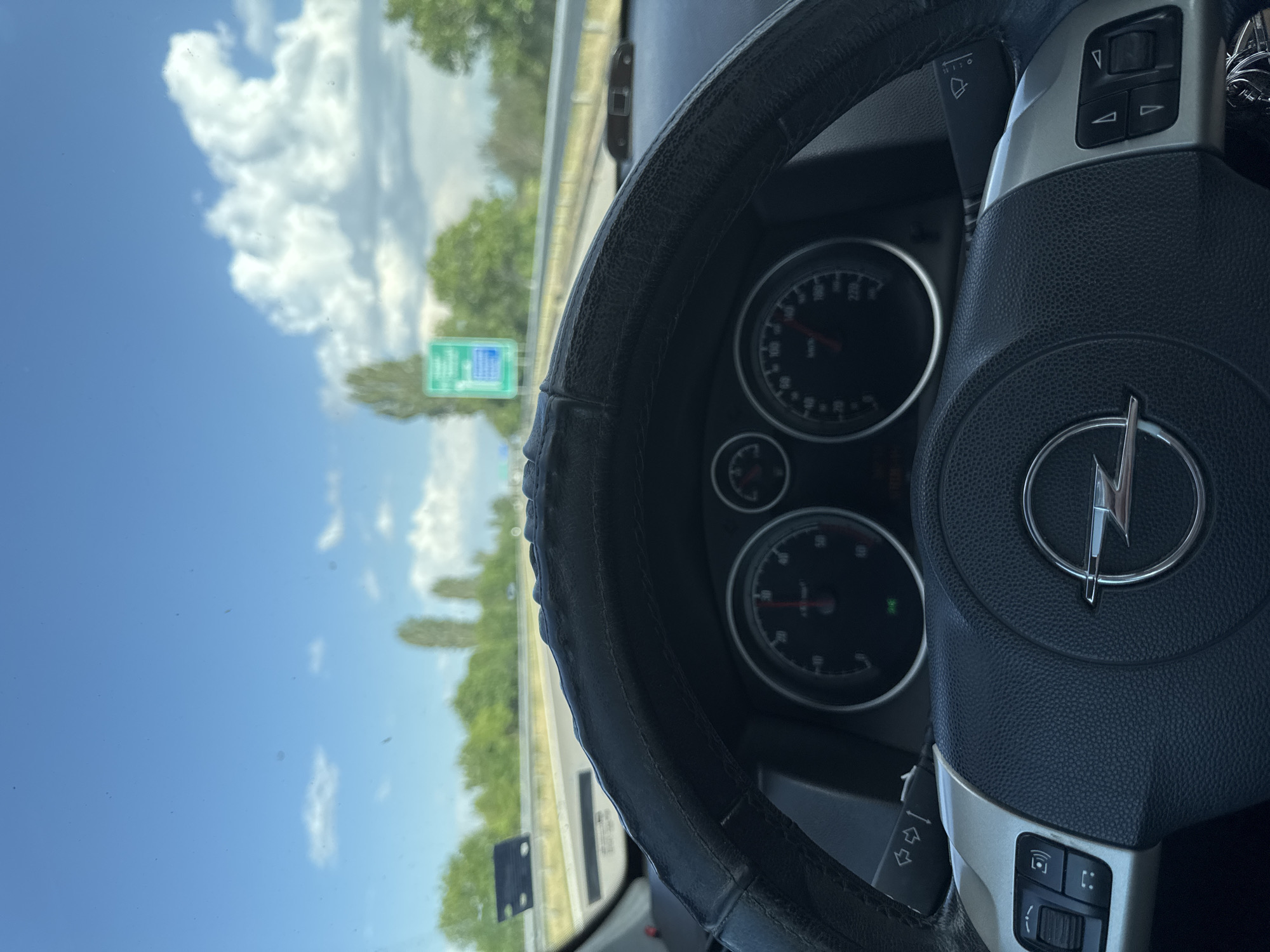 The image shows the interior of a car, specifically the steering wheel and dashboard. The car's speedometer and other gauges are visible. Through the windshield, a highway and sky with clouds are seen. A road sign is present on the side of the highway. Based on the road sign colors and steering wheel logo, the image seems to be taken while traveling near the city of Kalugerovo, Bulgaria.