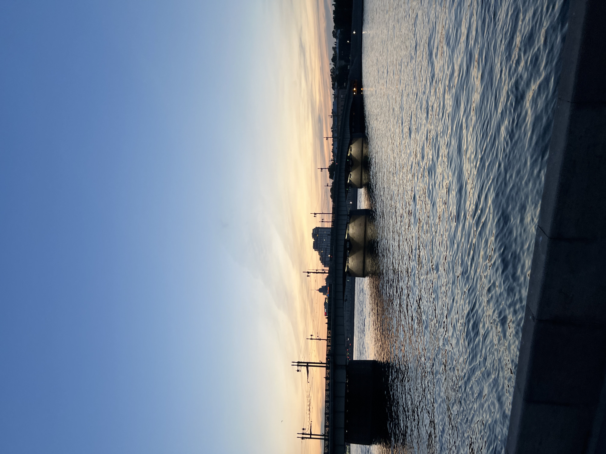 The image captures a wide shot of a bridge spanning across a body of water at dusk. The sky is a gradient of deep blue at the top, transitioning to pale yellow and orange hues near the horizon, indicating the presence of a sunset or sunrise. Wispy clouds are visible in the distance.
The bridge, constructed of concrete and steel, is supported by large pilings. It has lampposts lining its edges, some of which are illuminated, casting a warm glow on the water below. Silhouettes of buildings and structures can be seen on the far bank of the water, with a prominent domed building visible in the center. The water is calm with gentle ripples, reflecting the ambient light. In the foreground, a dark, concrete embankment is visible, suggesting a waterfront promenade or quay. The overall atmosphere is serene and tranquil, typical of an evening cityscape.