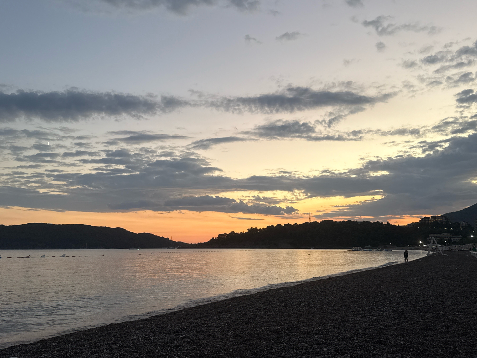 This is a wide shot of a pebble beach and the Adriatic Sea at dusk, located in Boreti, Montenegro. The sky is a gradient of pale blue at the top, transitioning to shades of orange and yellow near the horizon, with scattered grey clouds. The sea is calm, reflecting the sky's colors. In the distance, dark, silhouetted hills form the coastline. Along the shoreline, there are several small boats moored, and a few beach amenities like buoys and water attractions are visible. A single person is walking on the beach in the middle-right of the frame. The foreground is dominated by the dark, textured pebble beach.
