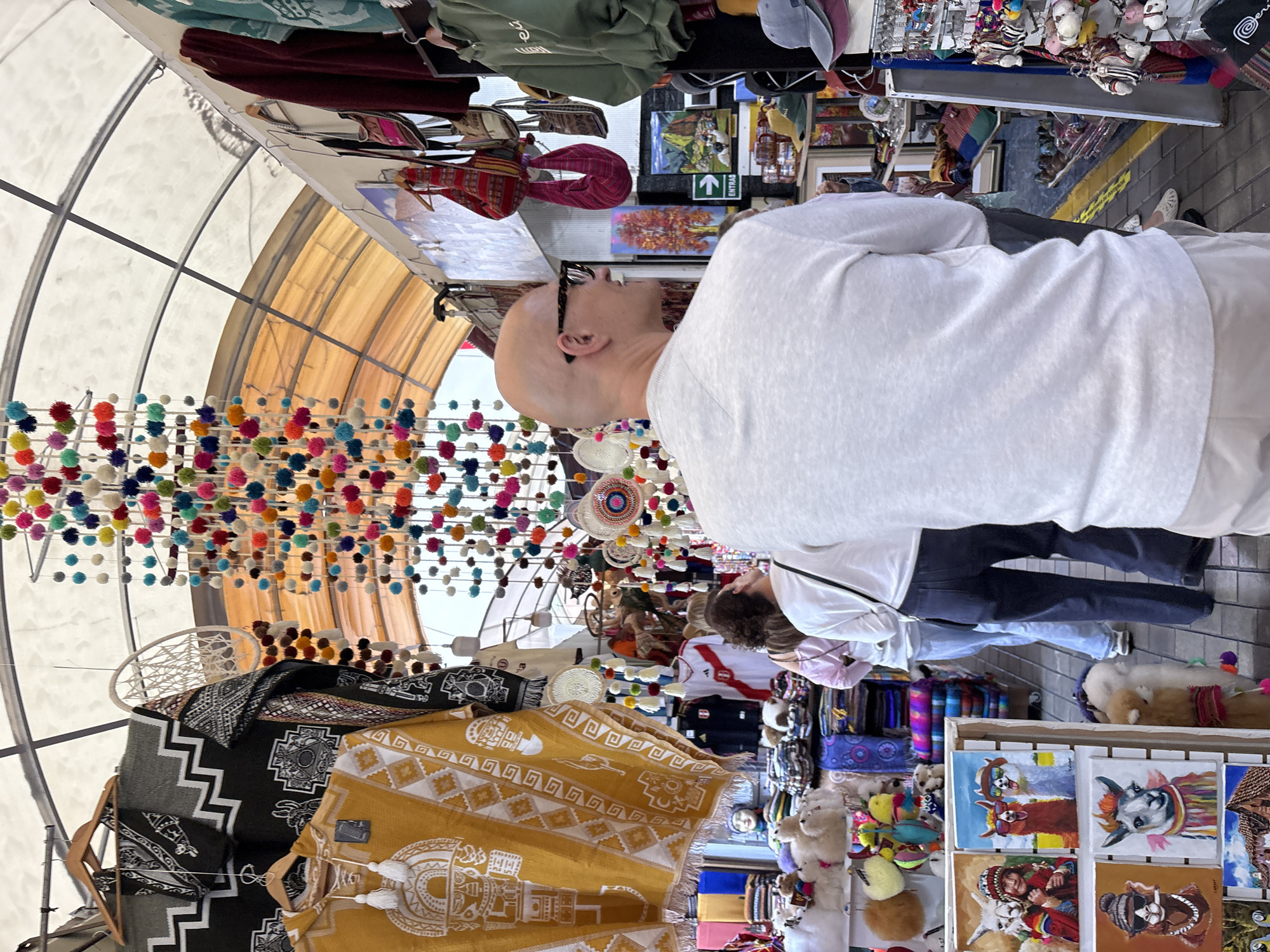 The image depicts a market stall in Miraflores, Peru, likely an artisan market, showcasing a variety of textiles and souvenirs. In the foreground, a bald man with glasses wearing a light-colored long-sleeved shirt and light pants faces away from the camera, looking towards the market stalls. To his left, two women are visible, one in a white long-sleeved shirt and blue jeans, and another in a pink top and blue jeans, both looking into the market.
The market stall features an assortment of items. Hanging from above is a colorful string of pom-poms in various hues, including red, yellow, blue, green, and pink. To the left, a large yellow poncho with white geometric patterns and figures is displayed, along with other dark-colored textiles with white designs. Further into the stall, rows of knitted items, possibly alpaca wool products, are arranged. Small paintings of llamas in different poses and with accessories are also visible.
The environment is an open-air market with a translucent roof supported by metal beams. The ground is paved with bricks. The overall impression is a vibrant and bustling market scene, typical of tourist areas in Peru.