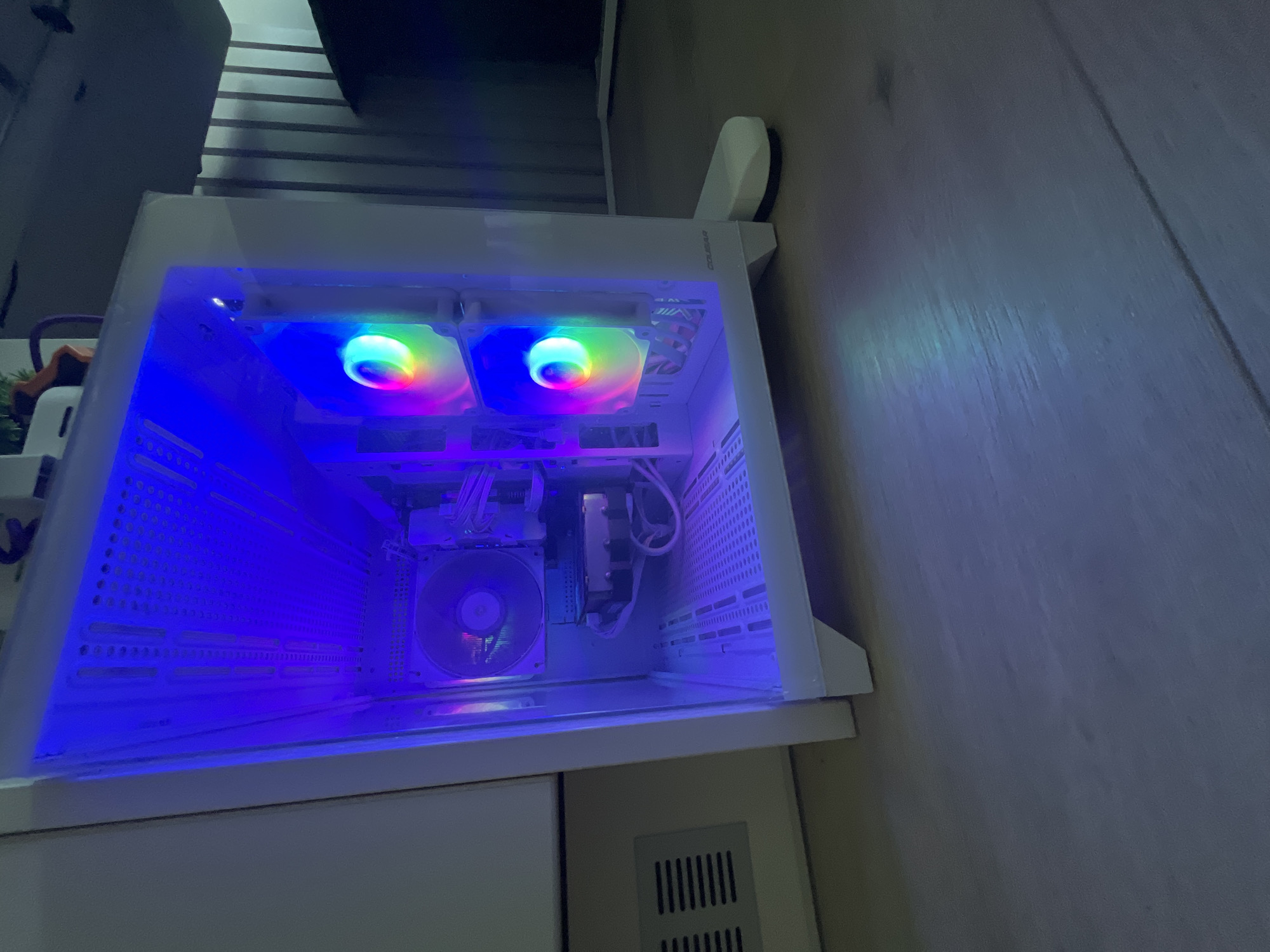 The image shows a desktop computer tower with transparent side panel. Inside, two rainbow-colored LED fans are visible at the top, and a third is visible at the back. The case is white. The computer is sitting on a white cabinet. The room appears to be indoors. The location is unknown; there is no indication of Varna, Bulgaria.