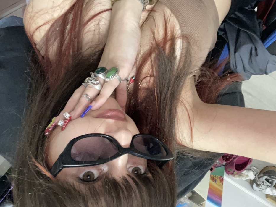 A woman wearing sunglasses and a brown top poses for the camera. Her hair is auburn and styled with bangs. She wears multiple rings on her fingers, including ones with green stones. Her nails are decorated with colorful designs. The background is cluttered with clothing and other objects. The overall perspective is an unusual angle, likely taken from above while she is lying down.