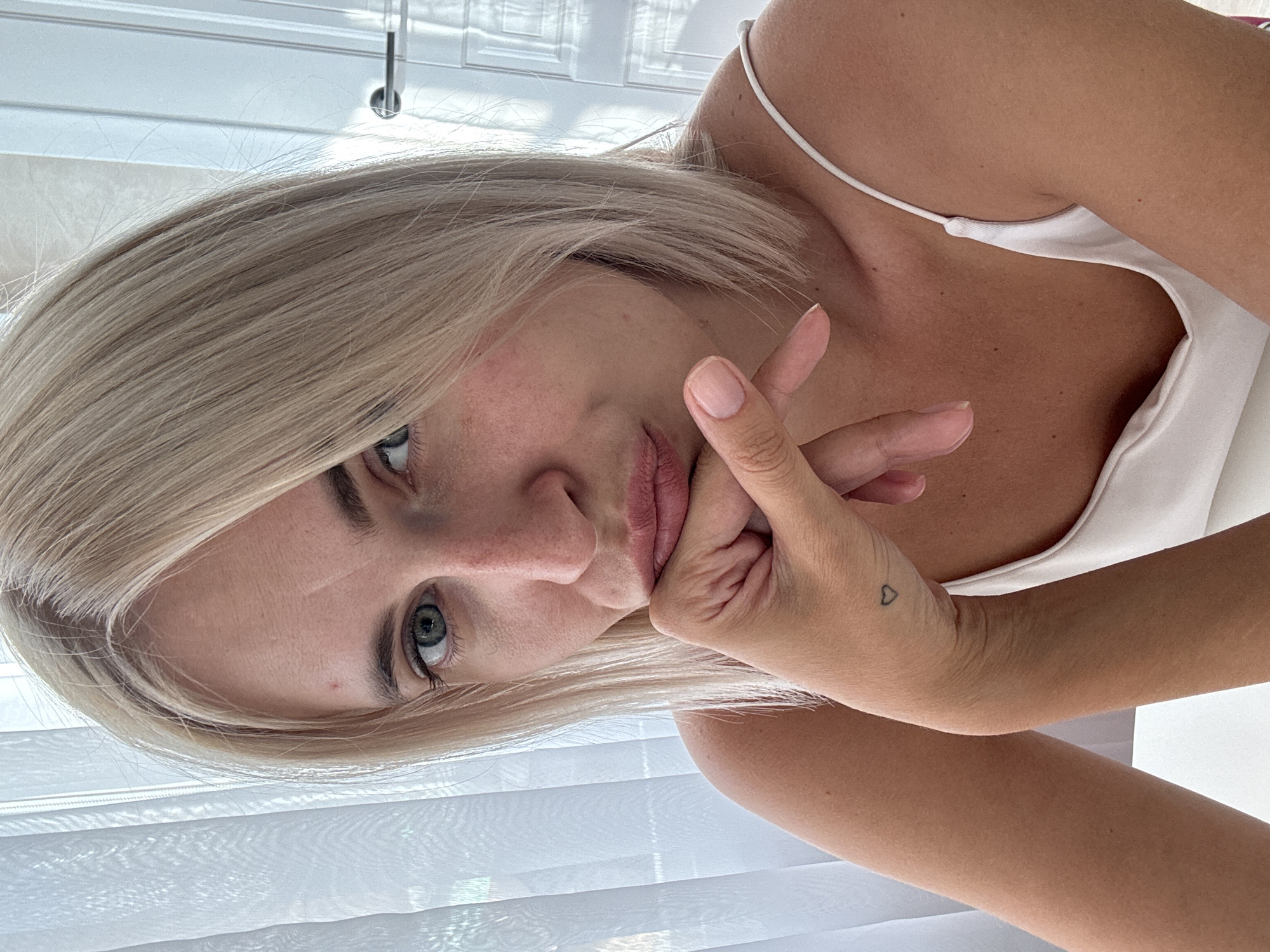 The media file shows a close-up, rotated portrait of a young woman with blonde hair. She is wearing a white camisole with a thin strap. Her left hand is resting on her chin, with her fingers touching her lips. A small, minimalist heart tattoo is visible on the back of her left hand. Her eyes are blue-green and she is looking directly at the camera with a neutral expression. The background appears to be indoors, with blinds providing a diffused light. The overall lighting suggests daytime. The image is oriented vertically, with the subject appearing to be lying down or leaning. The location context is Saratov, Russia.