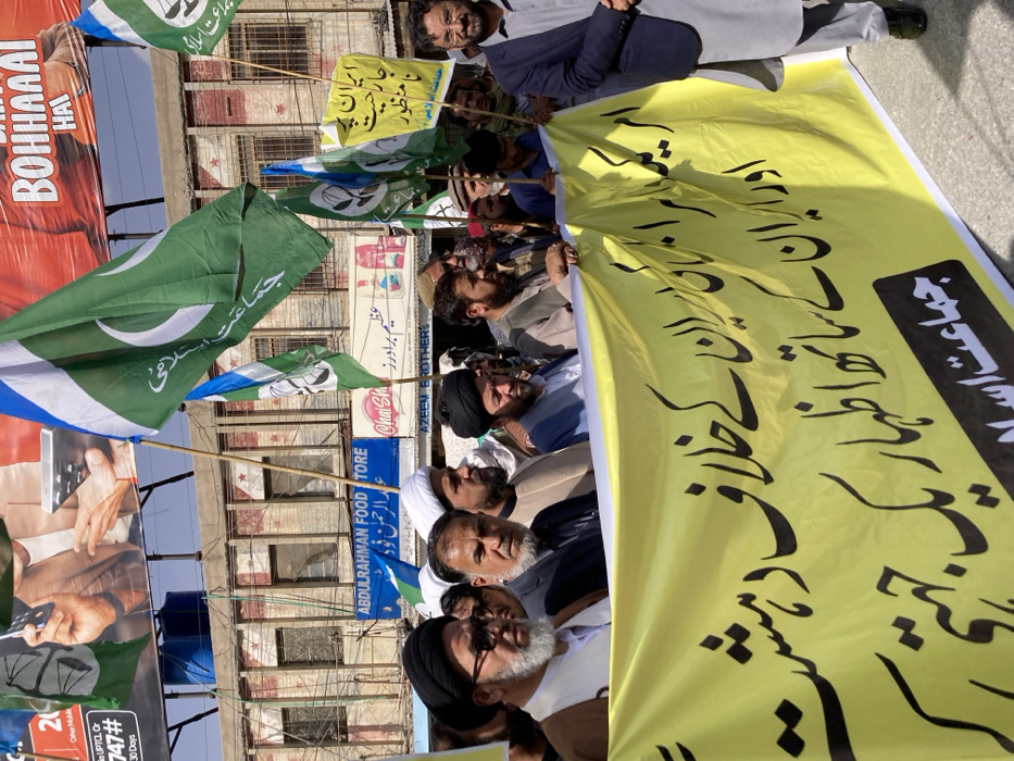 A street-level protest takes place on a clear day in Quetta City Tehsil, Pakistan. Approximately two dozen adult men are visible, marching or standing in a formation. In the foreground, a large yellow banner is horizontally held by multiple men. The banner features black Urdu text, reading: "امریکی اسرائیل کے خلاف دہشتگردی اور ایران کے ساتھ اظہار یکجہتی. جماعت اسلامی" (American-Israeli terrorism against Iran and solidarity with Iran. Jamaat-e-Islami).
Notable individuals in the front row, from left to right, include a man with a white beard, glasses, and a black turban; a man with a dark beard wearing a dark suit jacket; a man with a white turban and dark beard; and a man with a black turban and dark beard. On the far right, a man with a dark beard, glasses, and a dark suit jacket is partially visible, holding a walking stick.
Multiple individuals in the crowd are holding tall wooden poles topped with green, white, and blue flags. These flags bear the white crescent moon and star, similar to the Pakistani flag, but with a blue stripe at the top and the text "جماعت اسلامی" (Jamaat-e-Islami) in white on the green field. A smaller yellow sign is also visible in the middle-right, held by a man, displaying Urdu text: "ایران پر جارحیت نامنظور" (Aggression against Iran is unacceptable), also attributed to Jamaat-e-Islami.
The background features a multi-story building with multiple windows, some boarded, and overhead wiring. Several shop signs are visible on the ground floor, including "ABDULRAHMAN FOOD STORE," "Chai Shai," and "AZEEM BROTHERS," all in English and Urdu. Above the buildings, large billboards display advertisements, one with prominent red text reading "BOHAAAAI HAI." The sky is clear and blue.